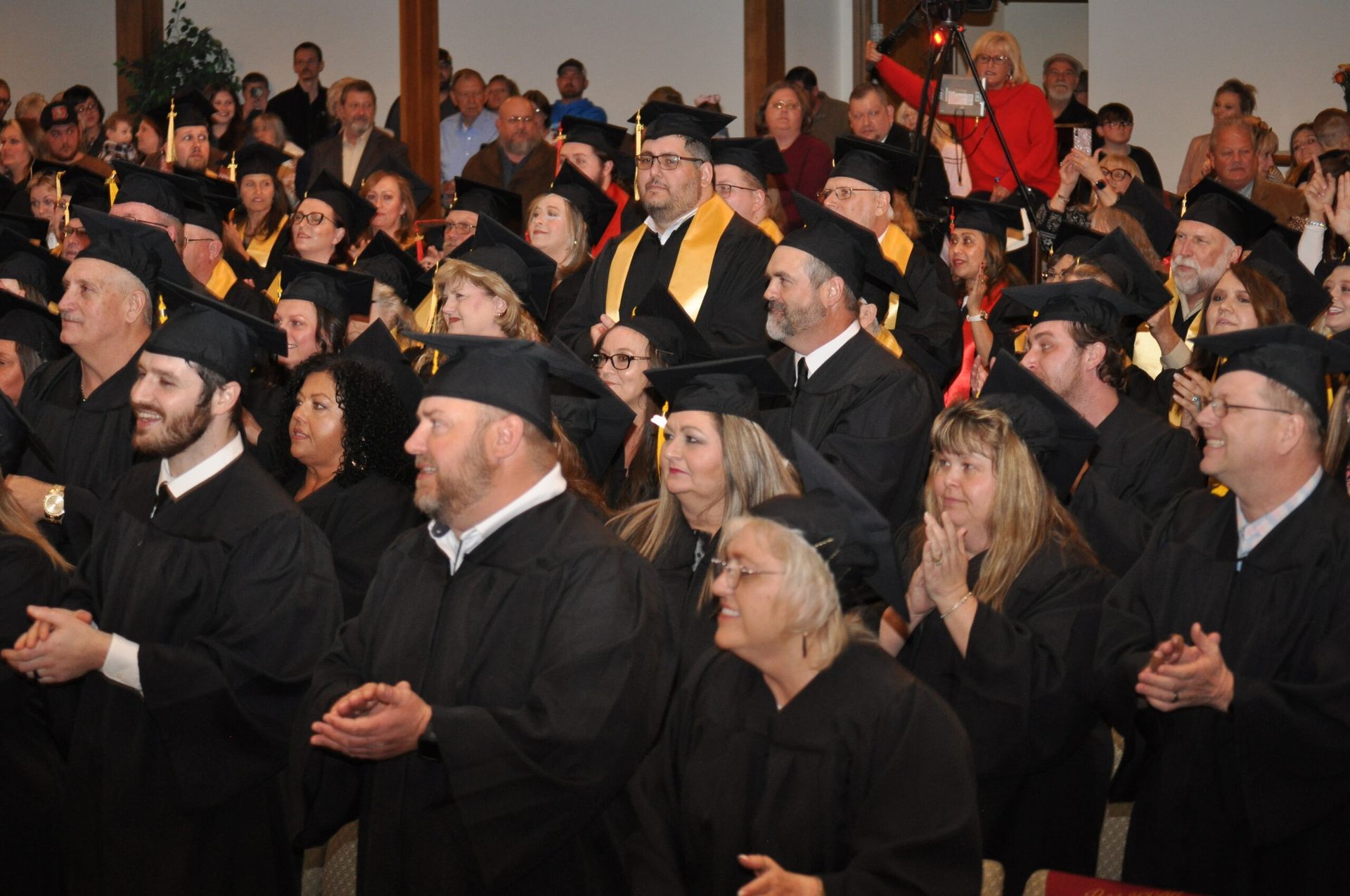 Graduation ceremony crowd clapping, wearing black gowns and mortarboards. Some have yellow stoles. Indoors, well-lit.
