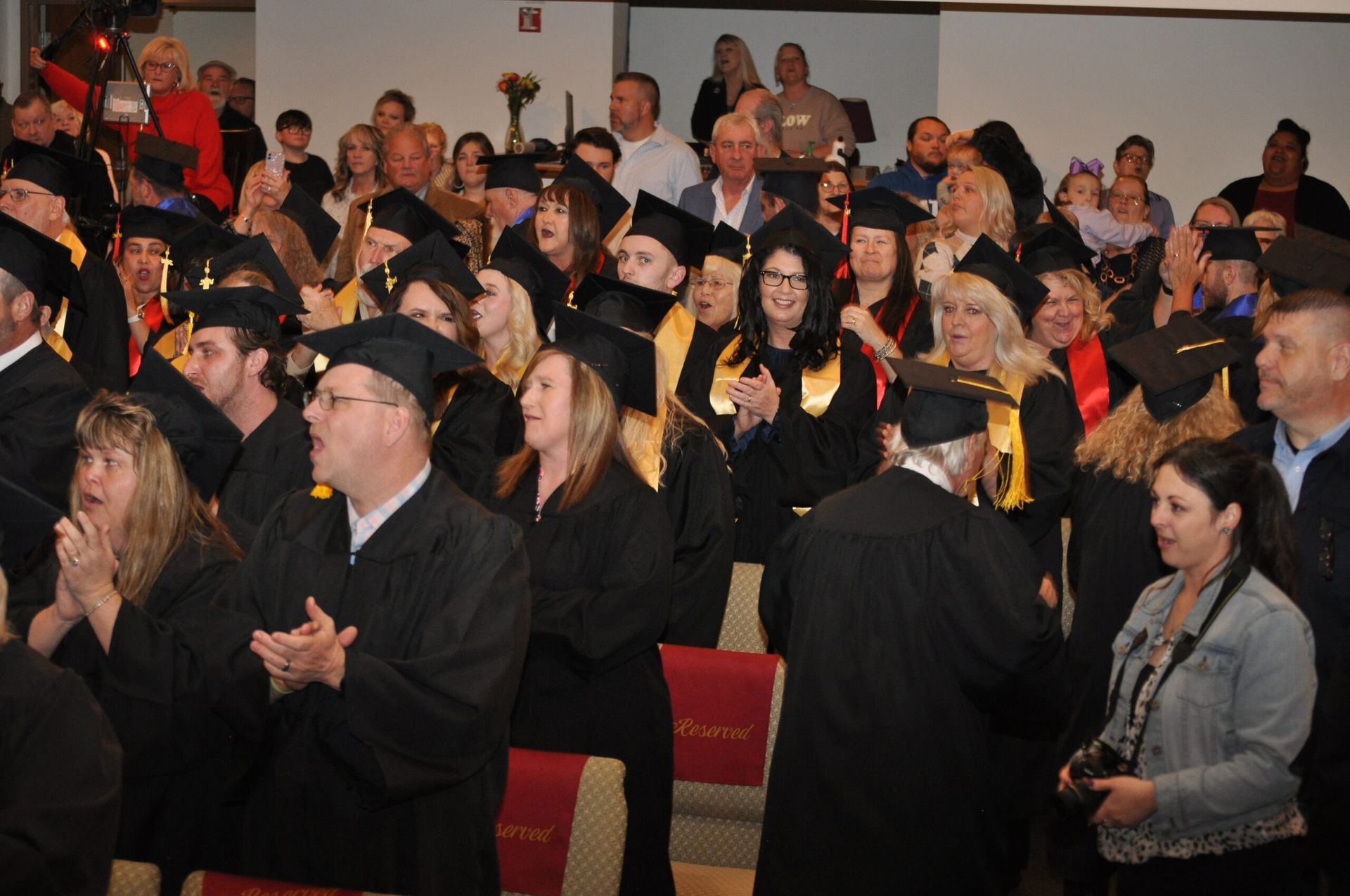 Graduates in black caps and gowns applaud at a ceremony, seated in rows, with an audience behind them.