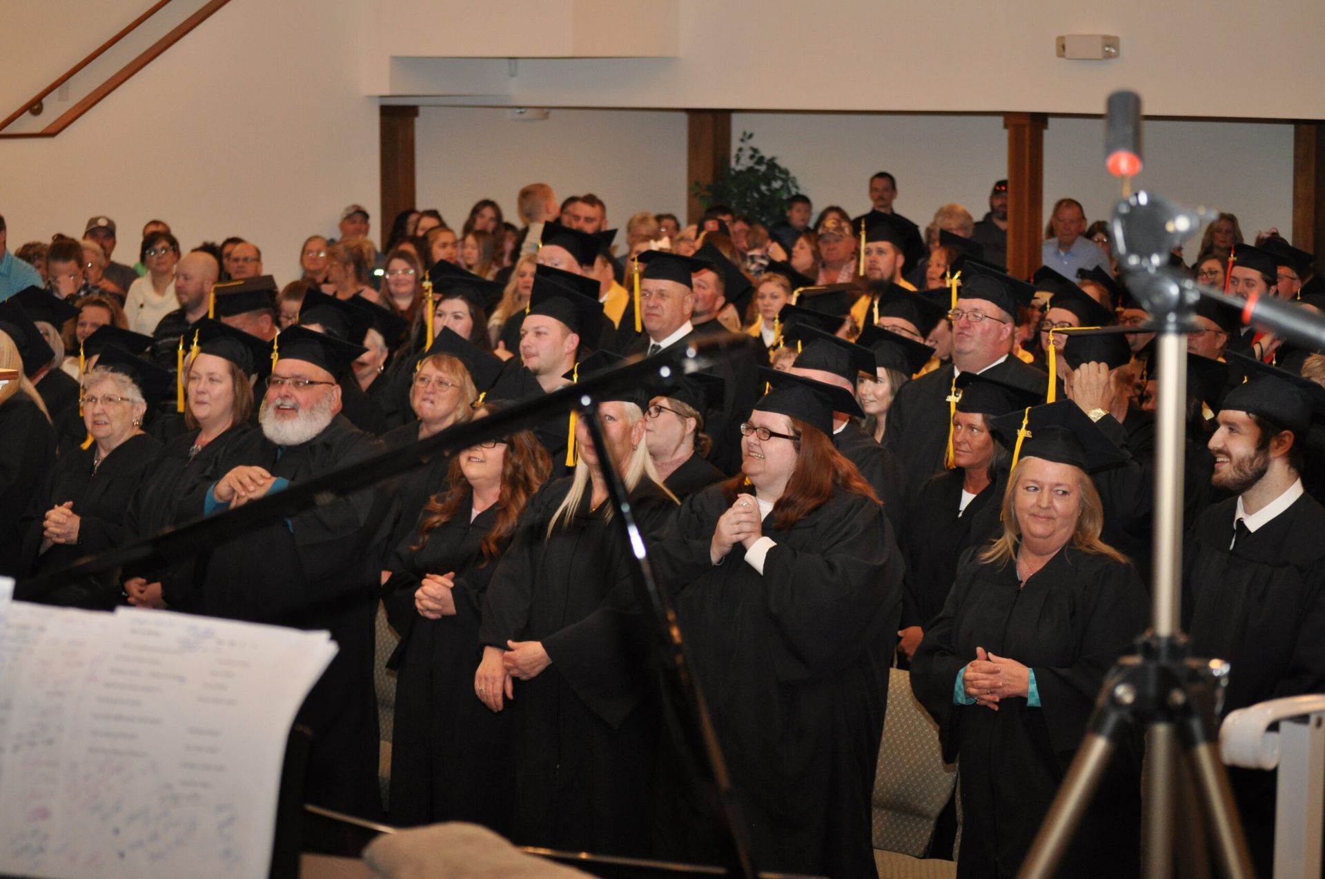 Graduation ceremony with graduates in black gowns and caps, audience in background, microphone on tripod.