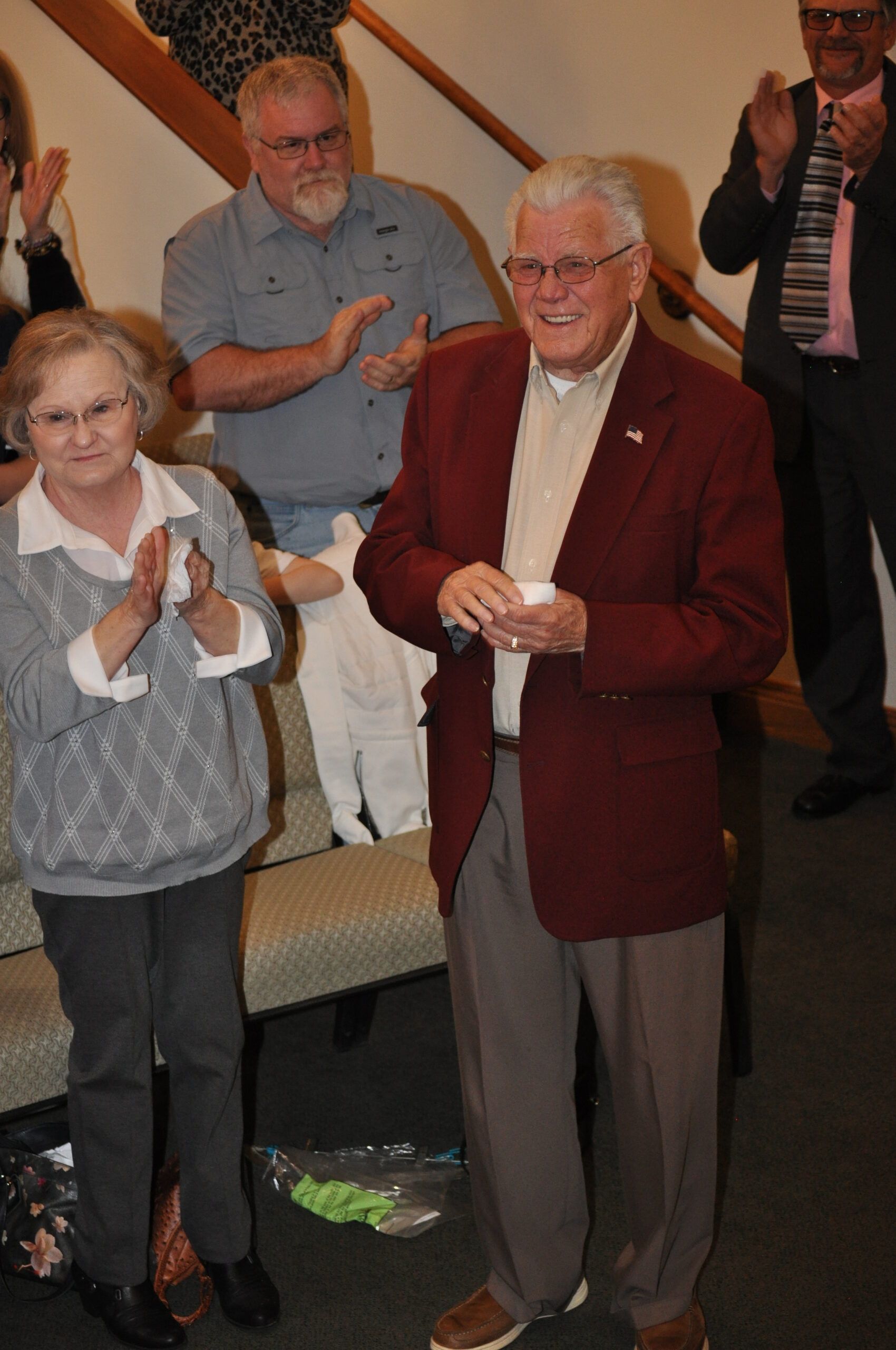 Man in red blazer smiles, applauded by people. Interior, near stairs.