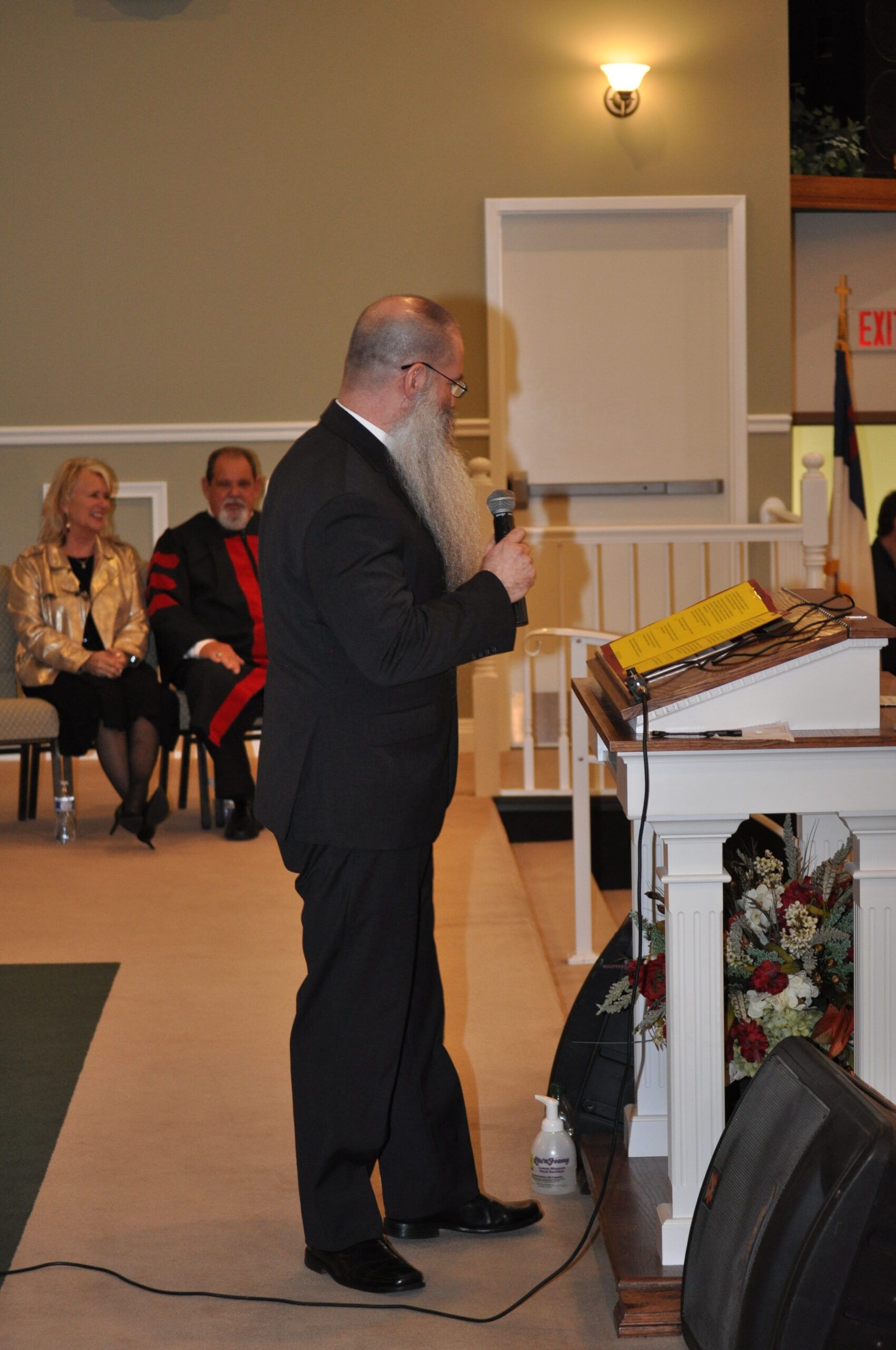 Man with long beard speaking at a podium; two people sit behind him.