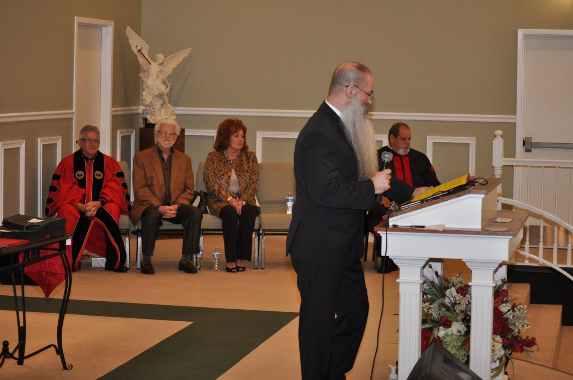 Man speaking at a podium with others seated behind. Ceremony setting, light colored room.