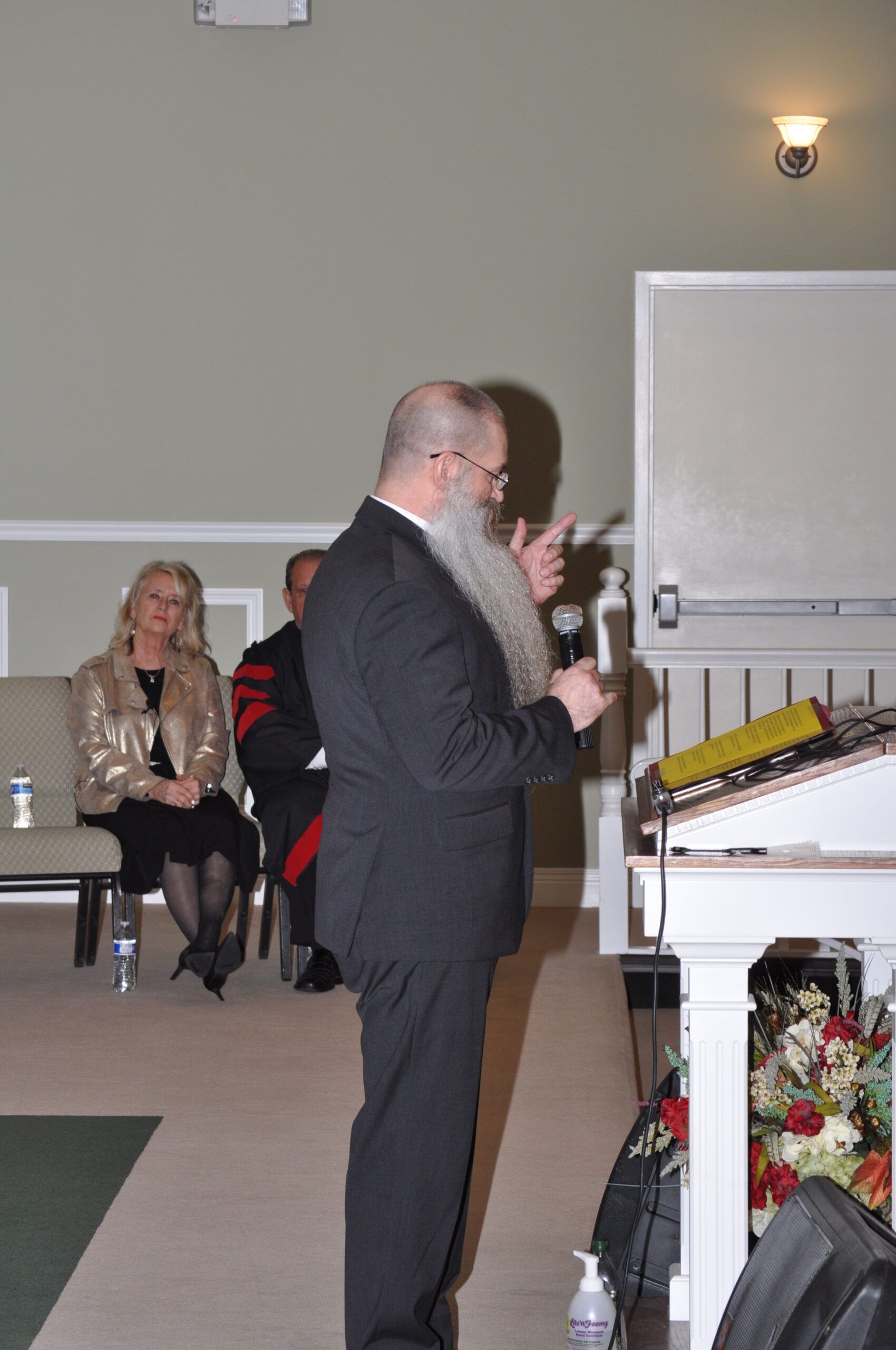 Man with long beard speaks at podium, other people in background.