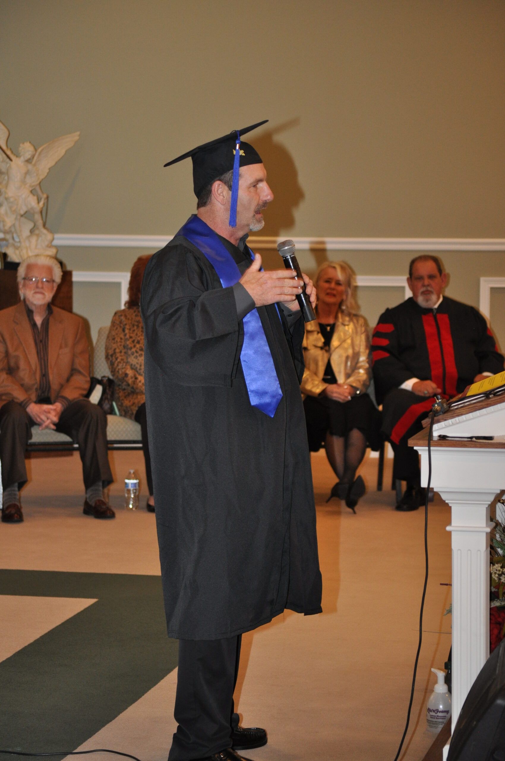 Man in graduation gown, speaking into a microphone. Formal setting with seated audience members.