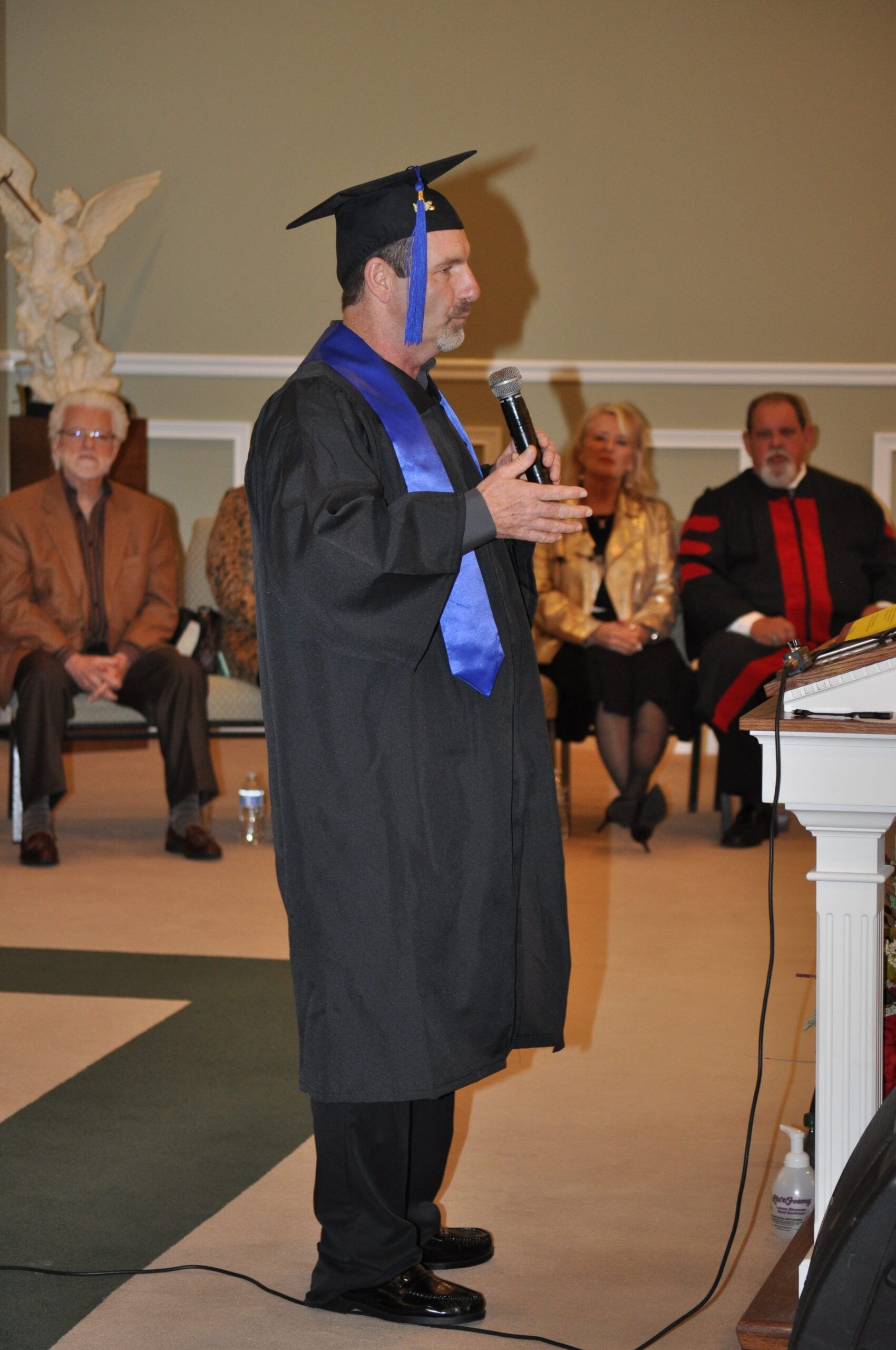 Man in graduation attire speaks at podium; others seated on stage.