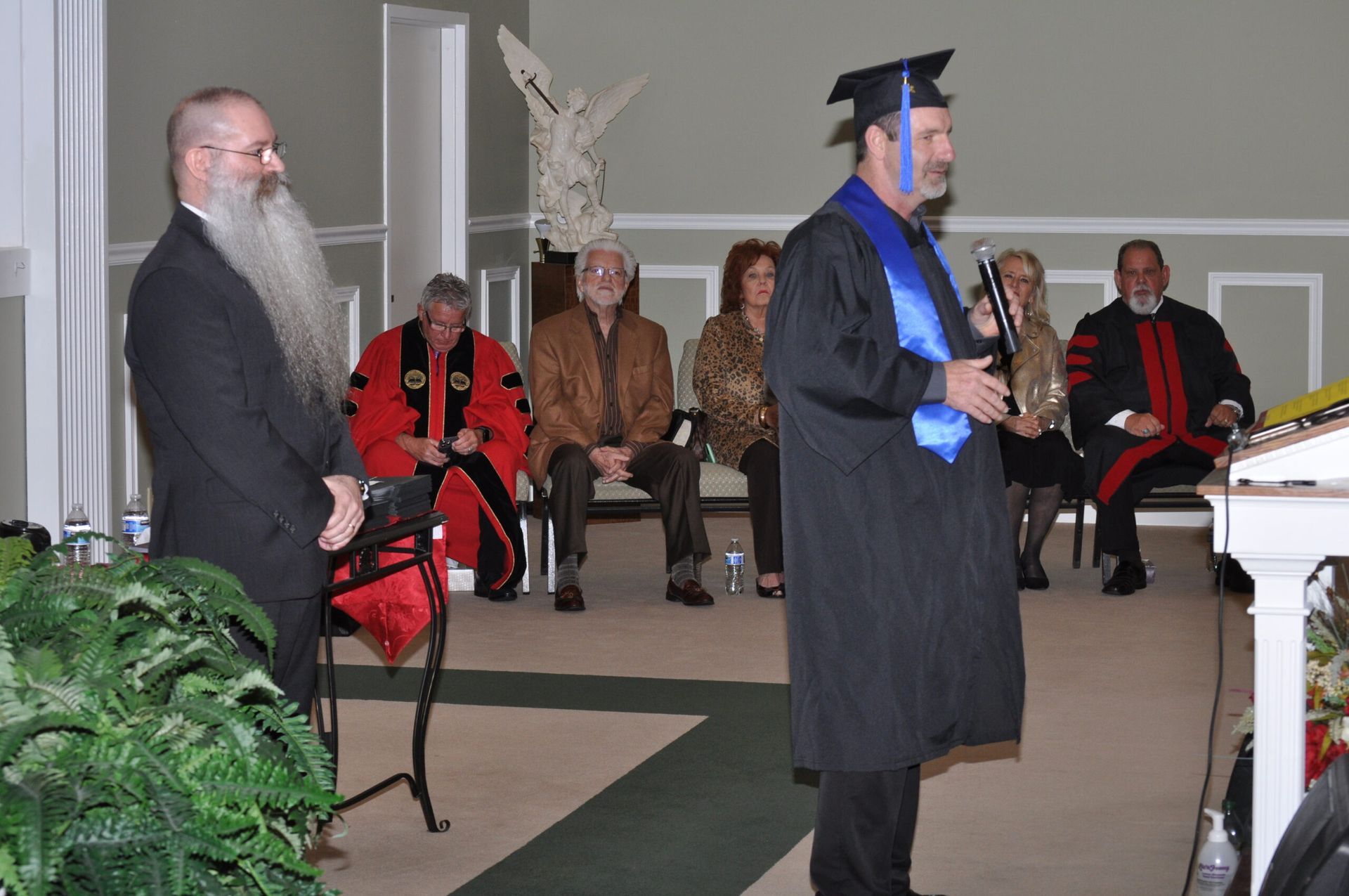 Man in graduation gown speaking at podium; audience seated, long-bearded man standing.