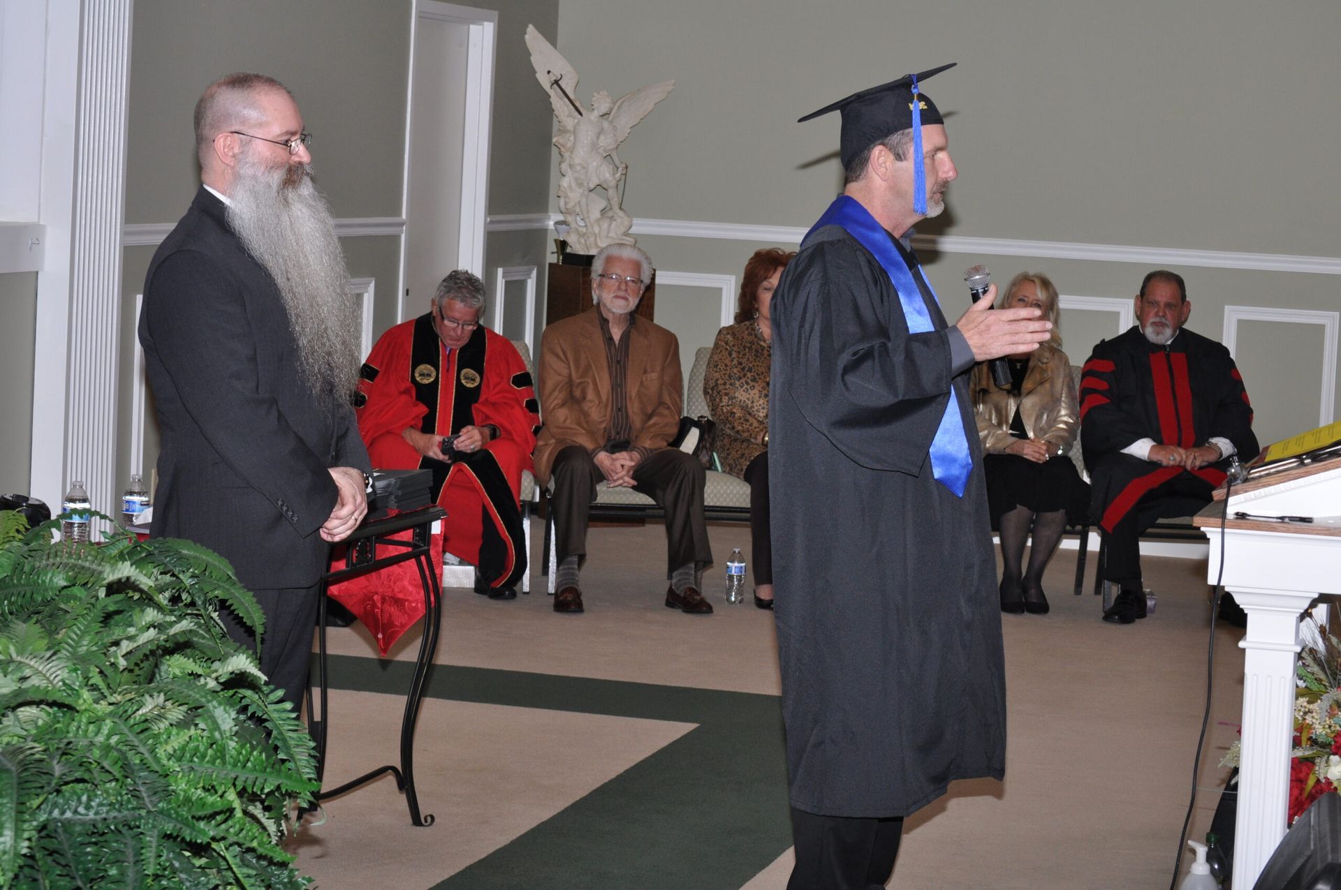 A man in a graduation gown speaks, standing at a podium. Others are seated in a chapel-like setting.