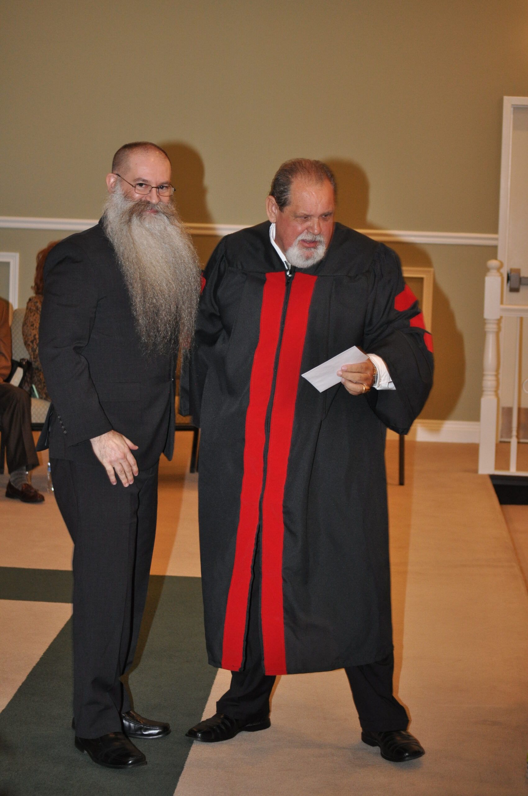Two men standing; one in a black robe with red trim, the other with a long beard. Indoors, likely ceremony.