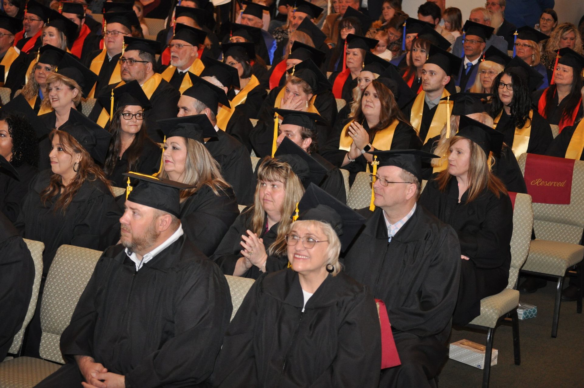 Graduates in black caps and gowns at a ceremony, many with yellow accents, seated in a hall.