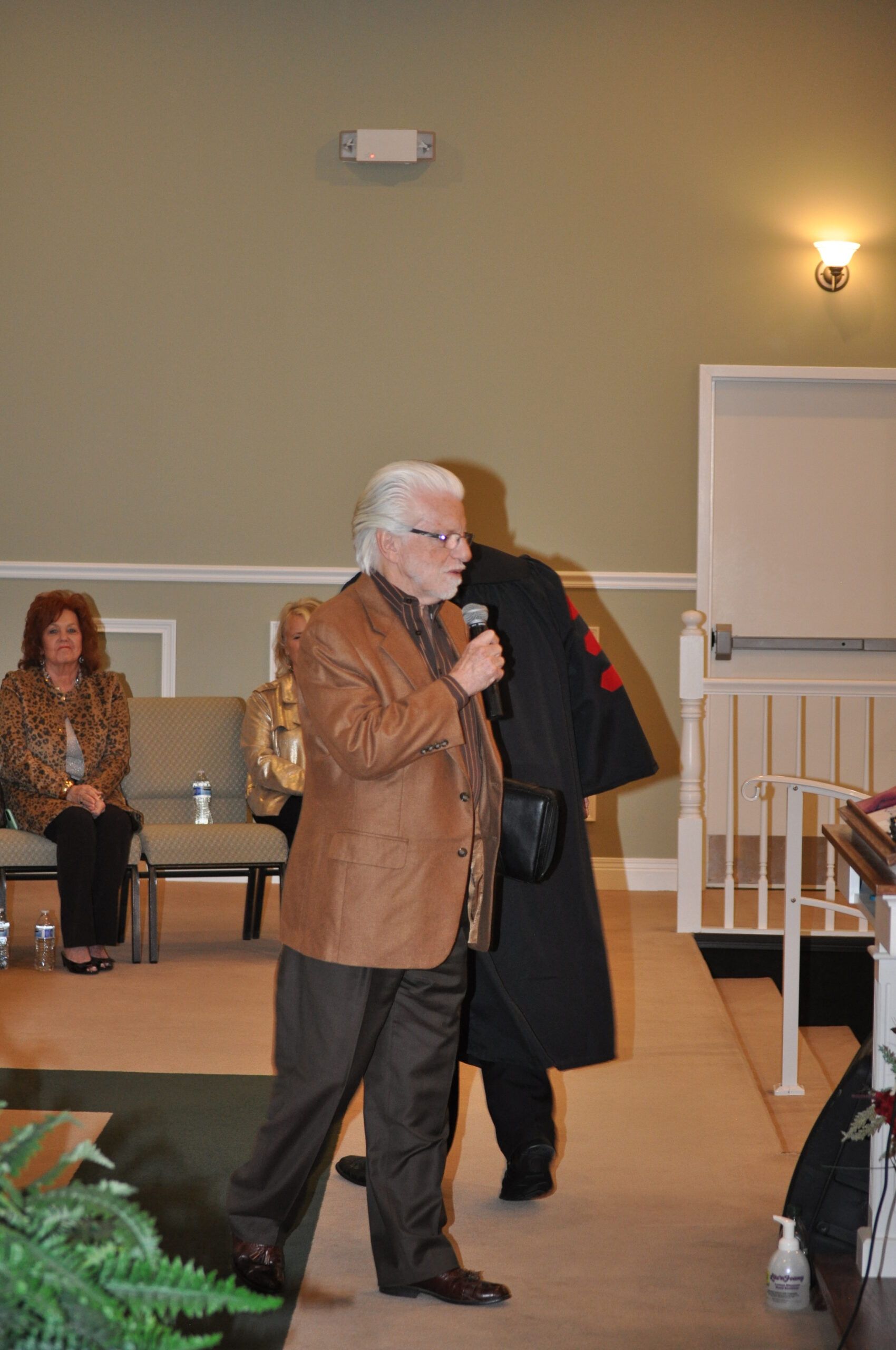 Man in brown jacket speaks into a microphone in a church; another person in black robe stands behind him.