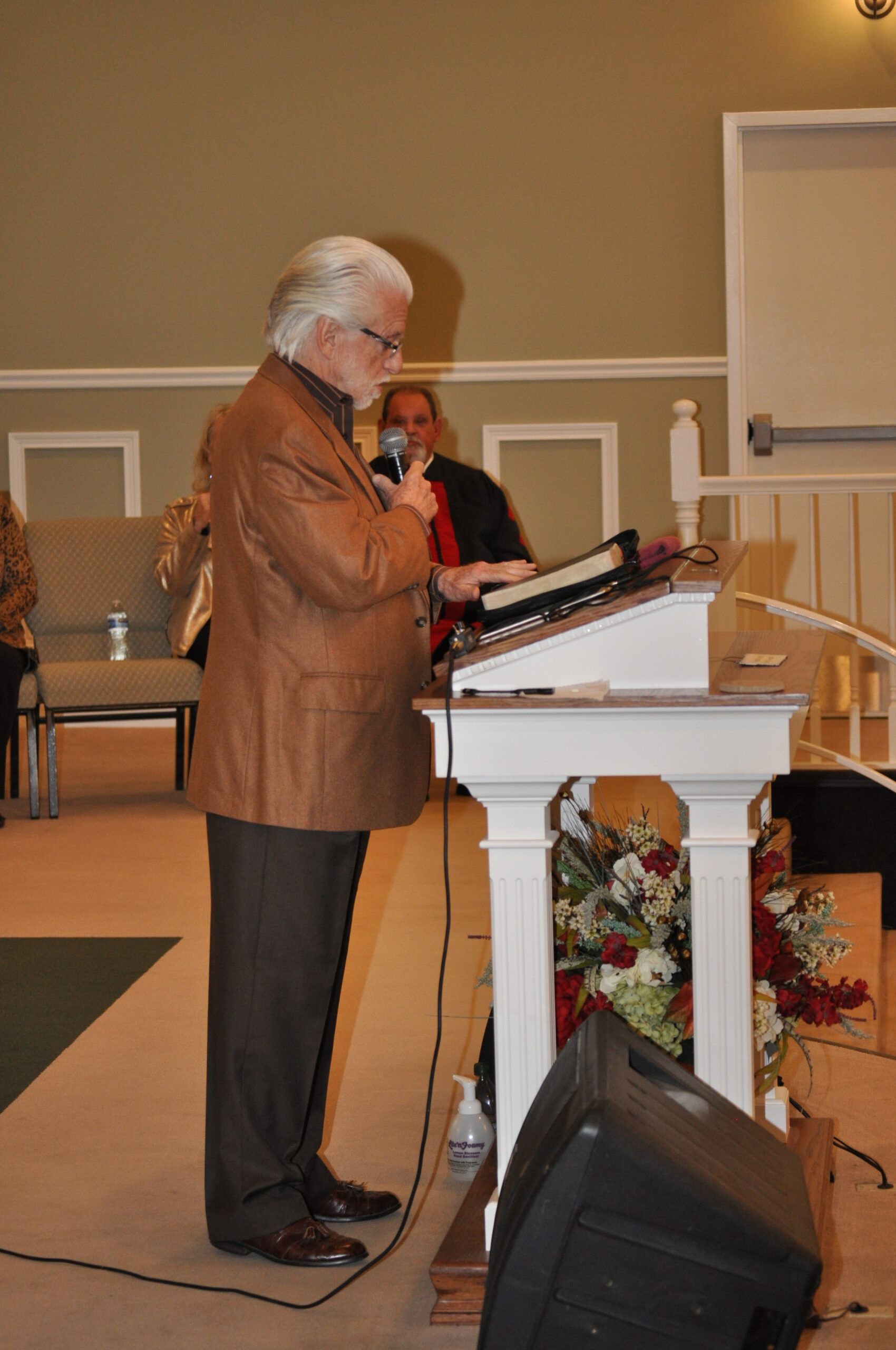 Man in brown jacket speaks at a podium; keyboard and sound equipment nearby. Church setting.