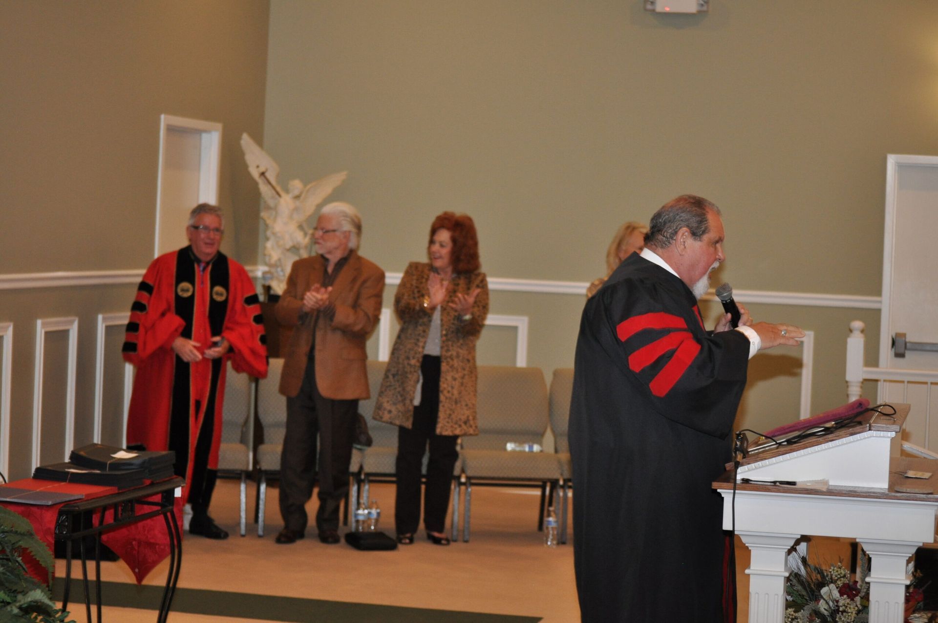 People in academic robes at a podium, clapping. Interior, likely a church or hall.