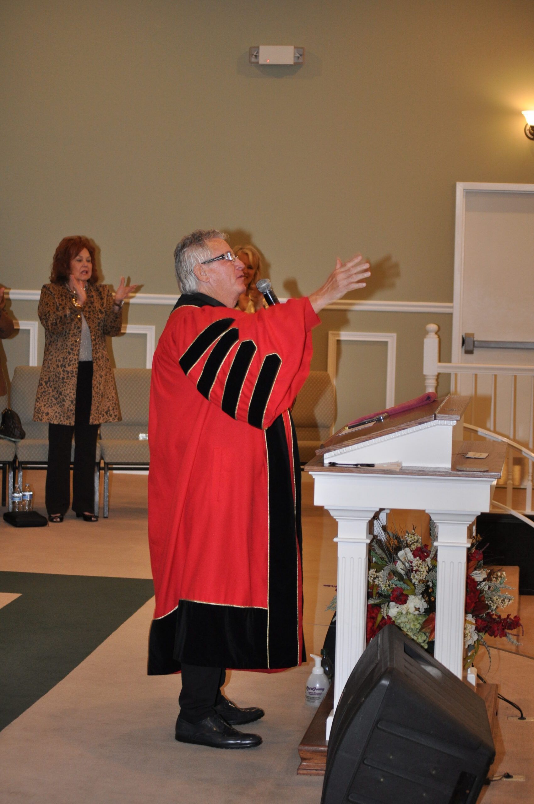 Person in red robe speaking at a lectern, arms outstretched; another person applauds in background.