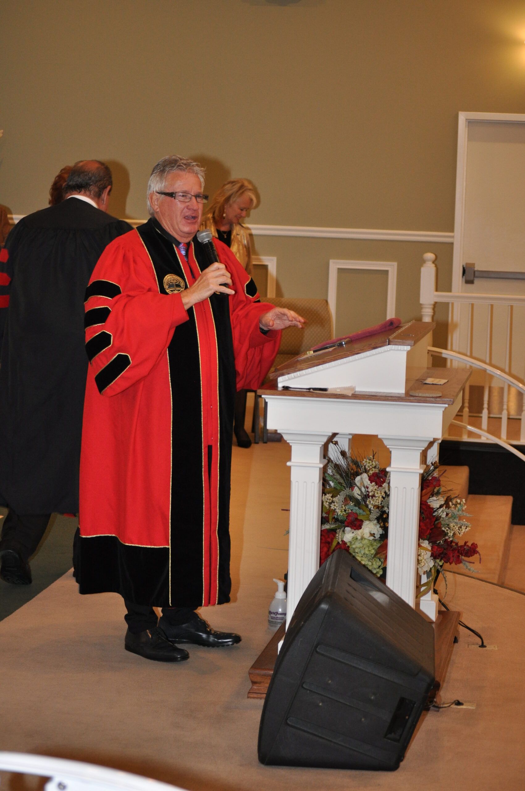 Man in red academic gown speaks at a lectern, holding a microphone.