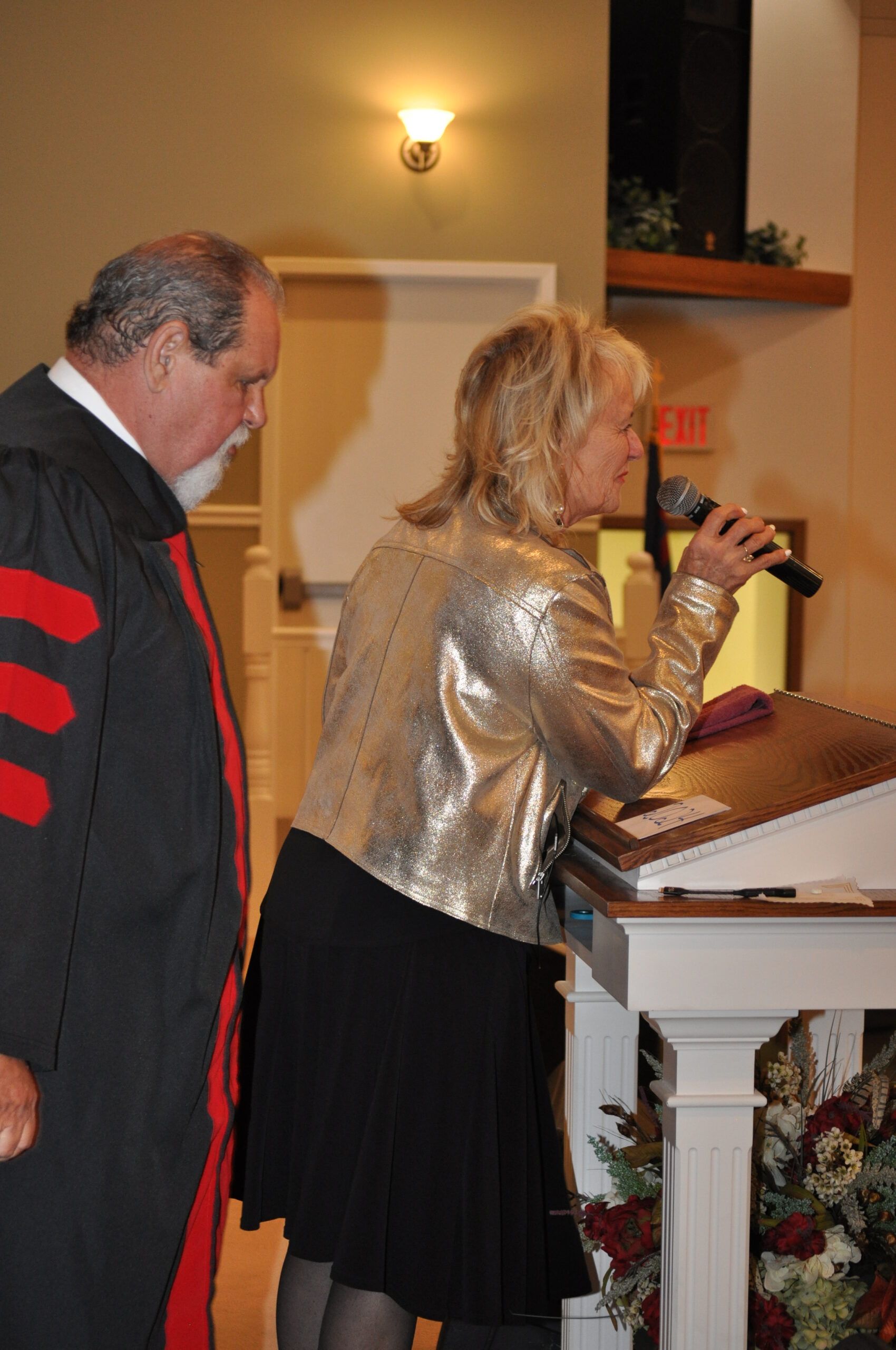 Woman in sequined jacket speaks at a podium; man in academic robe stands beside her. Indoor setting.