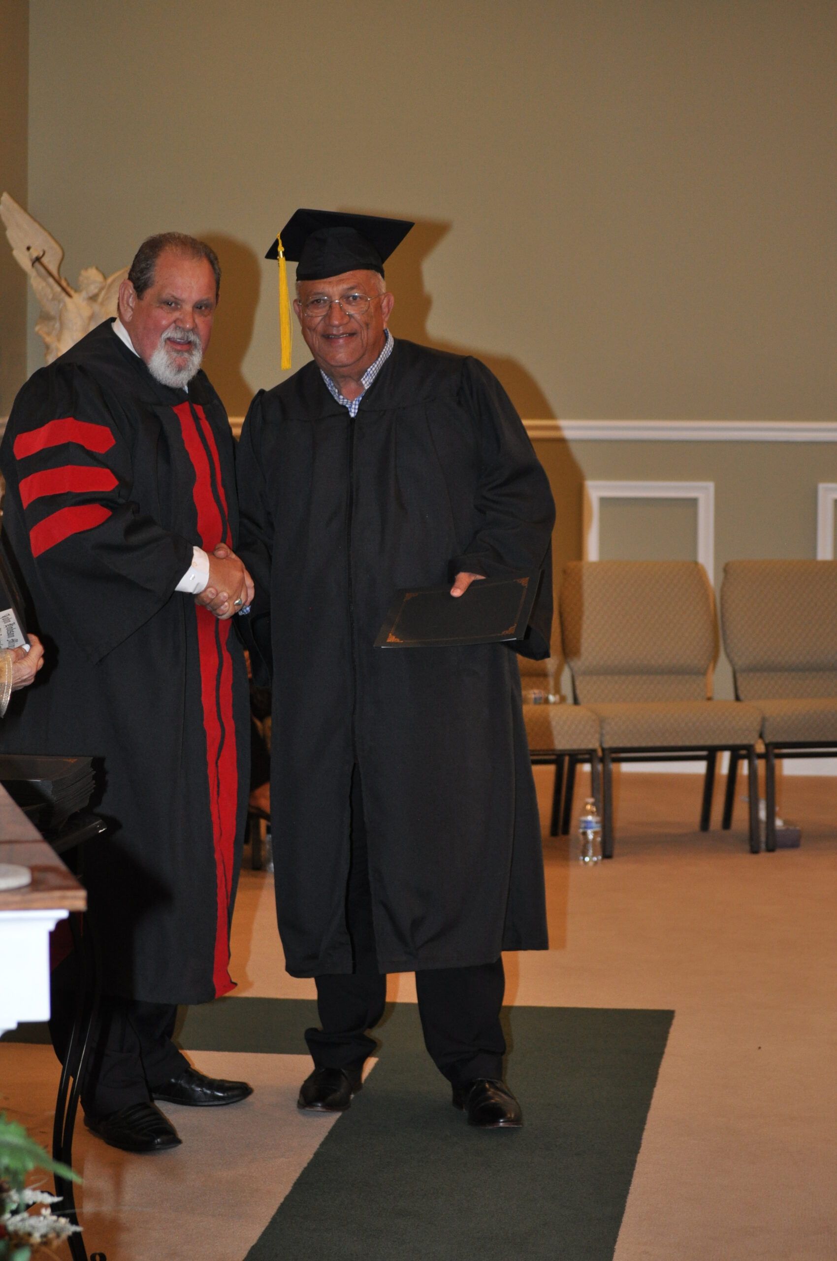 Man in graduation gown receiving diploma from another man. Indoors, church-like setting.