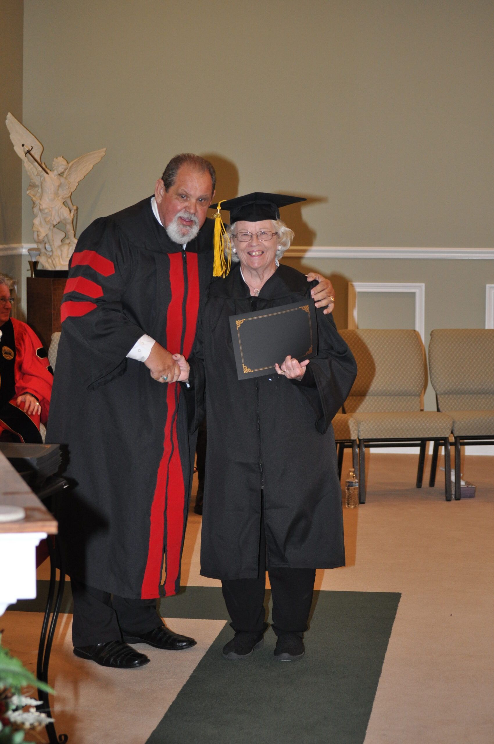 Man in academic robe shakes hands with graduate wearing cap and gown; holding a diploma.