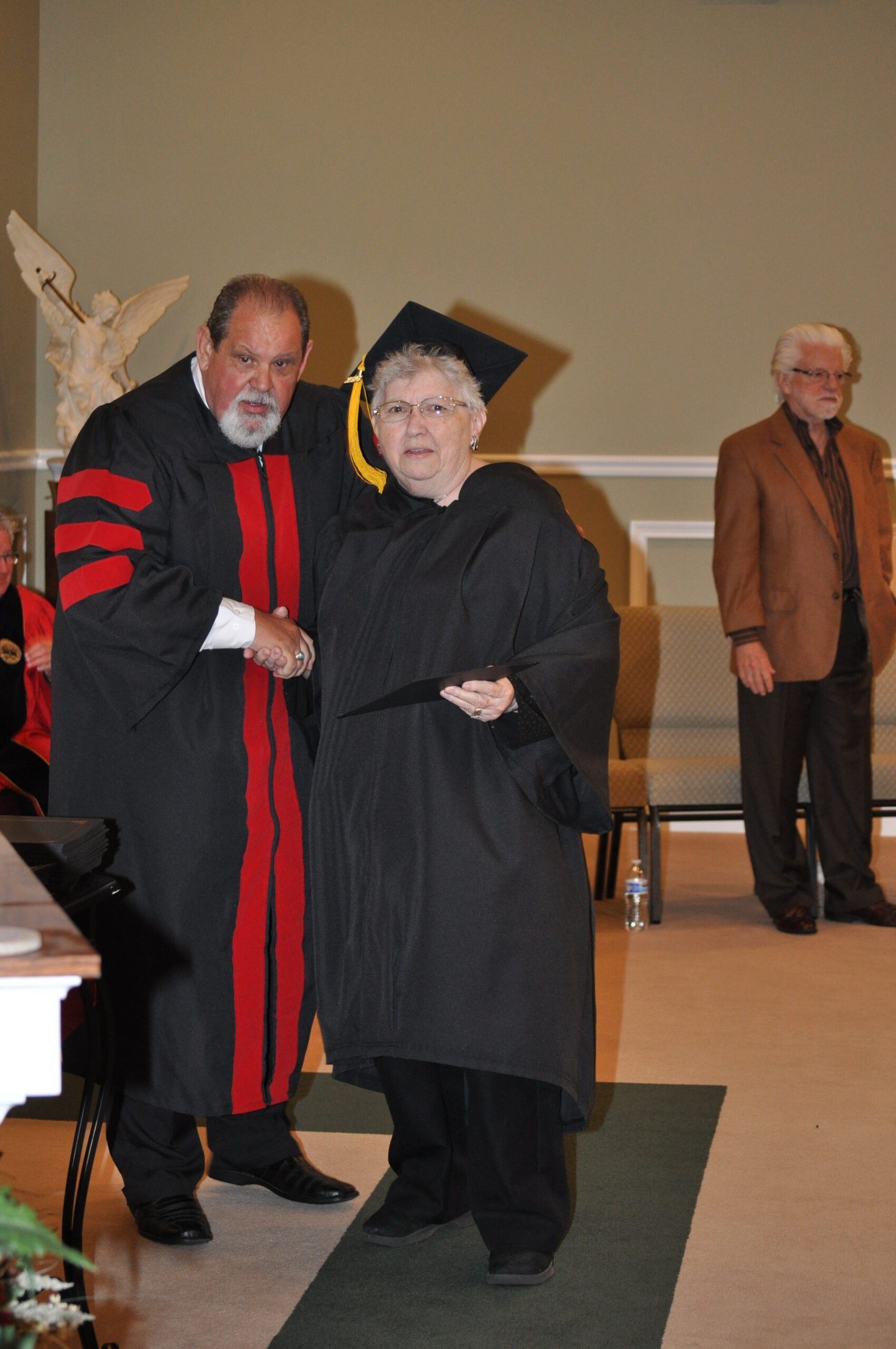 Person in graduation gown receiving diploma from person in academic robe.