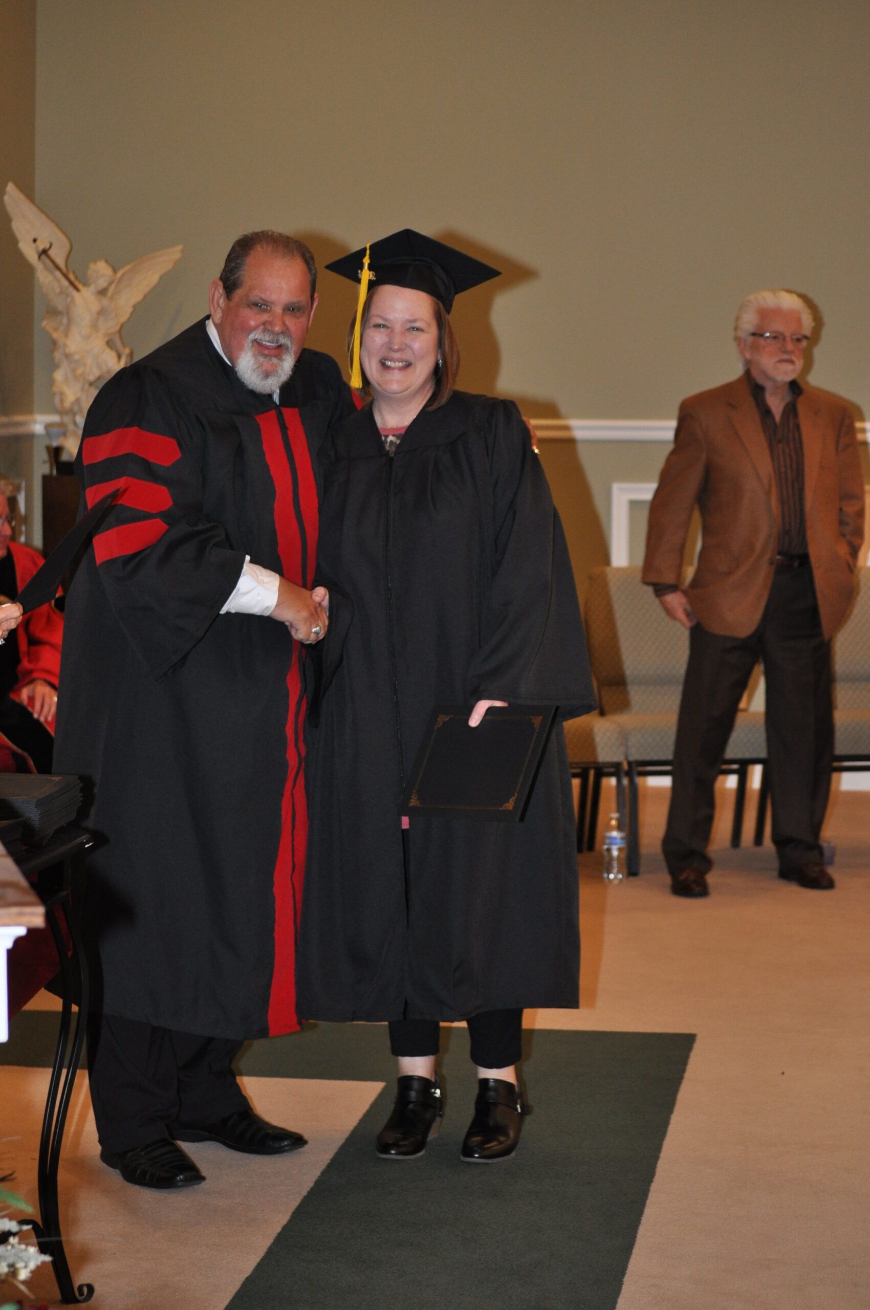 Woman in graduation gown shaking hands with robed man on stage; another man watches.