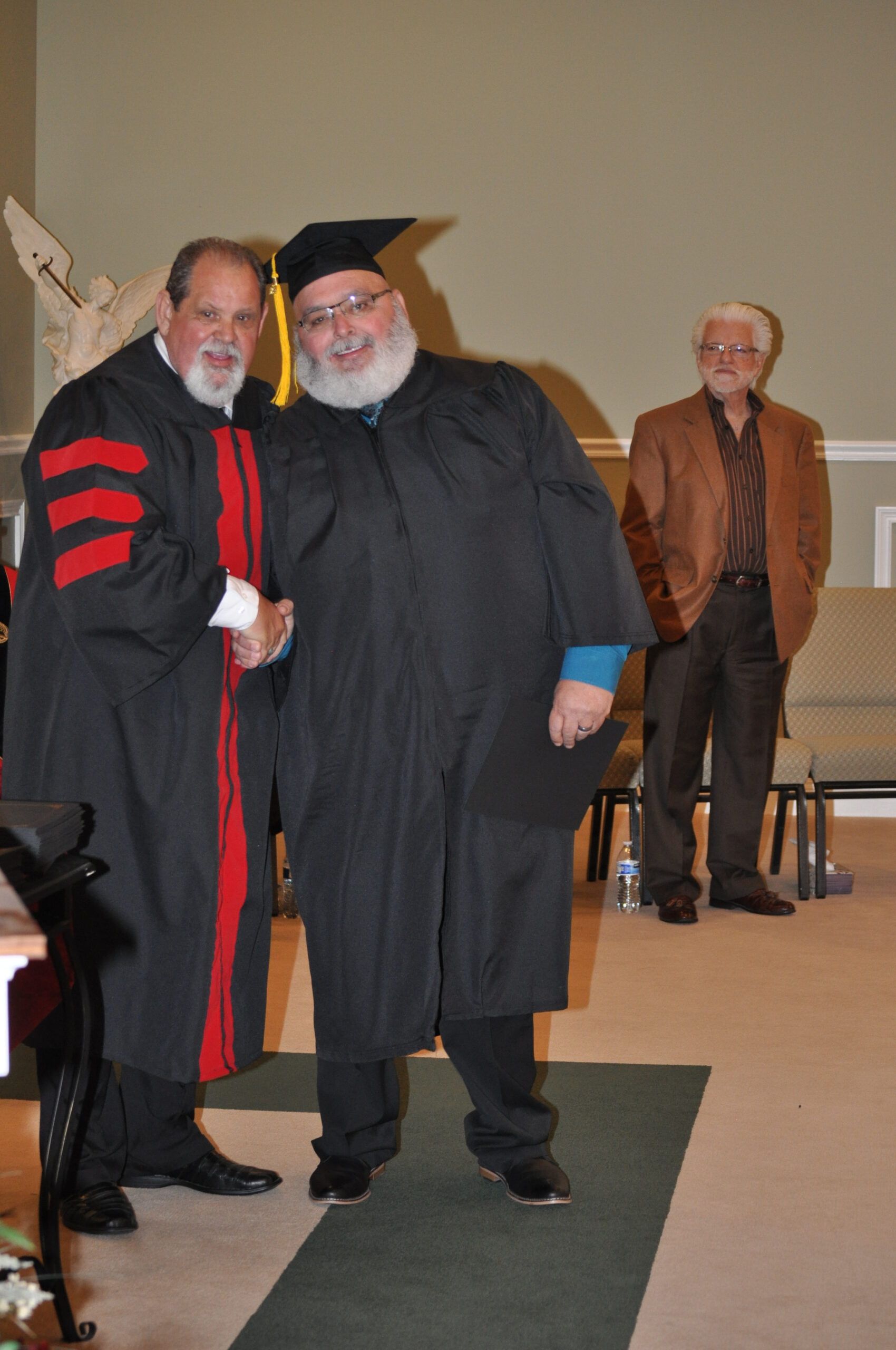 Man in graduation gown shakes hands with a man in academic regalia, a third man watches in background.