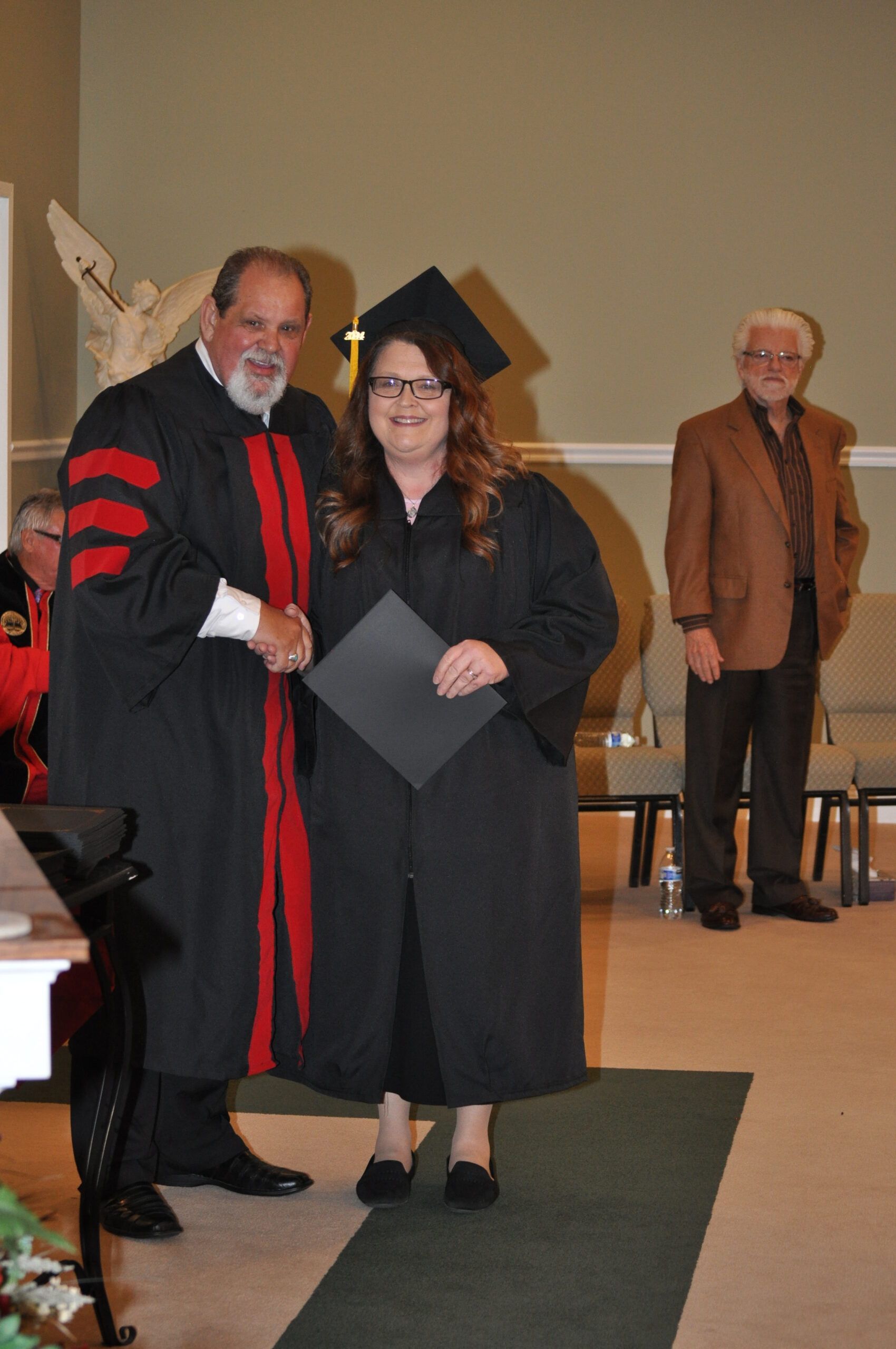 A woman in cap and gown shakes hands with a man in academic robes after receiving a diploma in an auditorium.