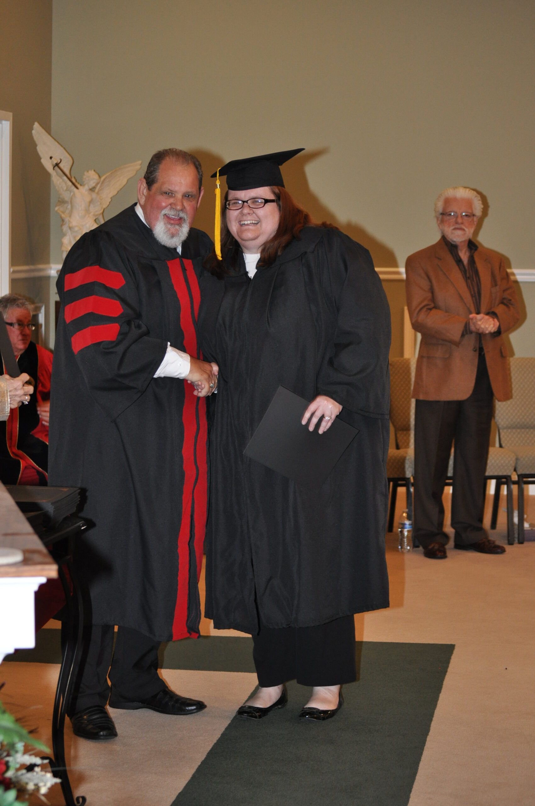 Person in graduation gown shakes hands with another person in formal attire; ceremony setting.
