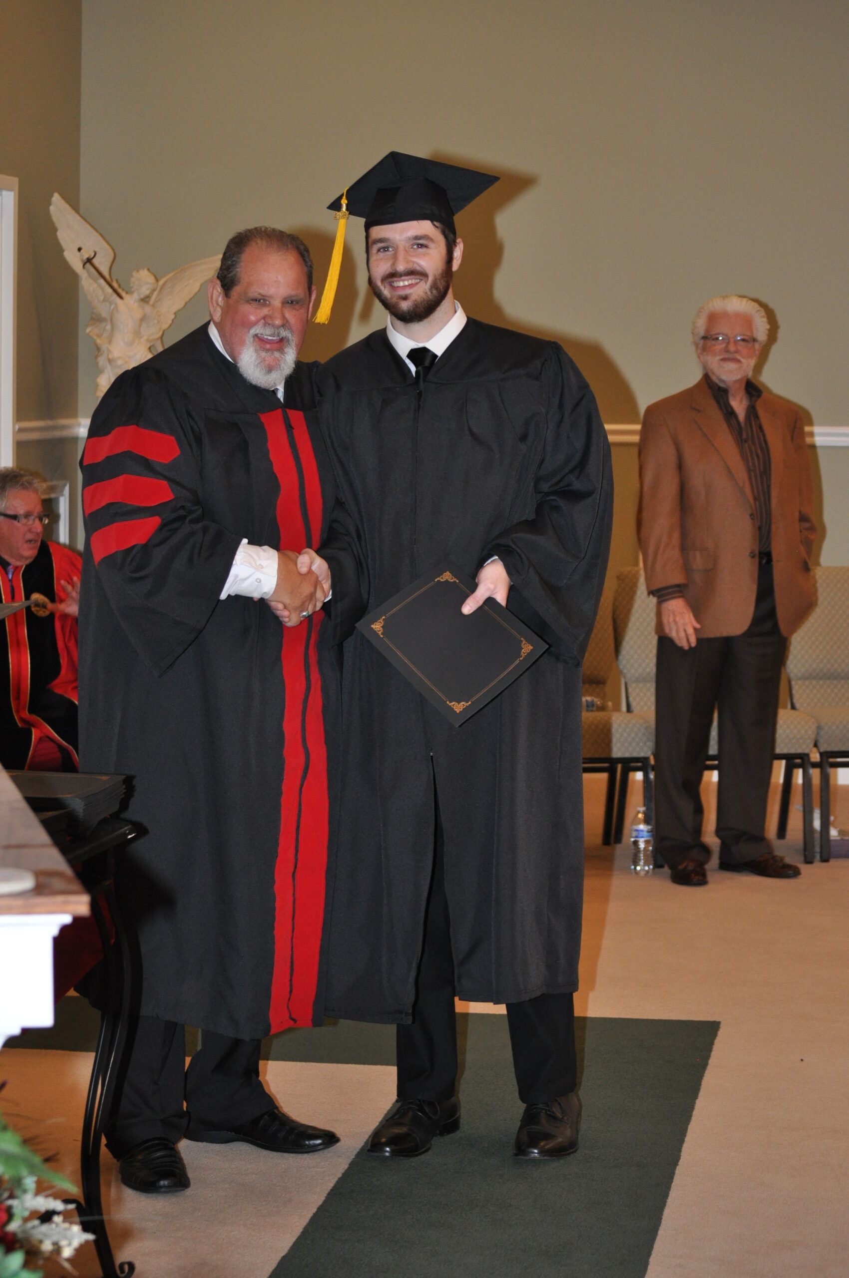 Man in graduation gown shakes hands with a man in academic regalia, holding a diploma, inside a building.