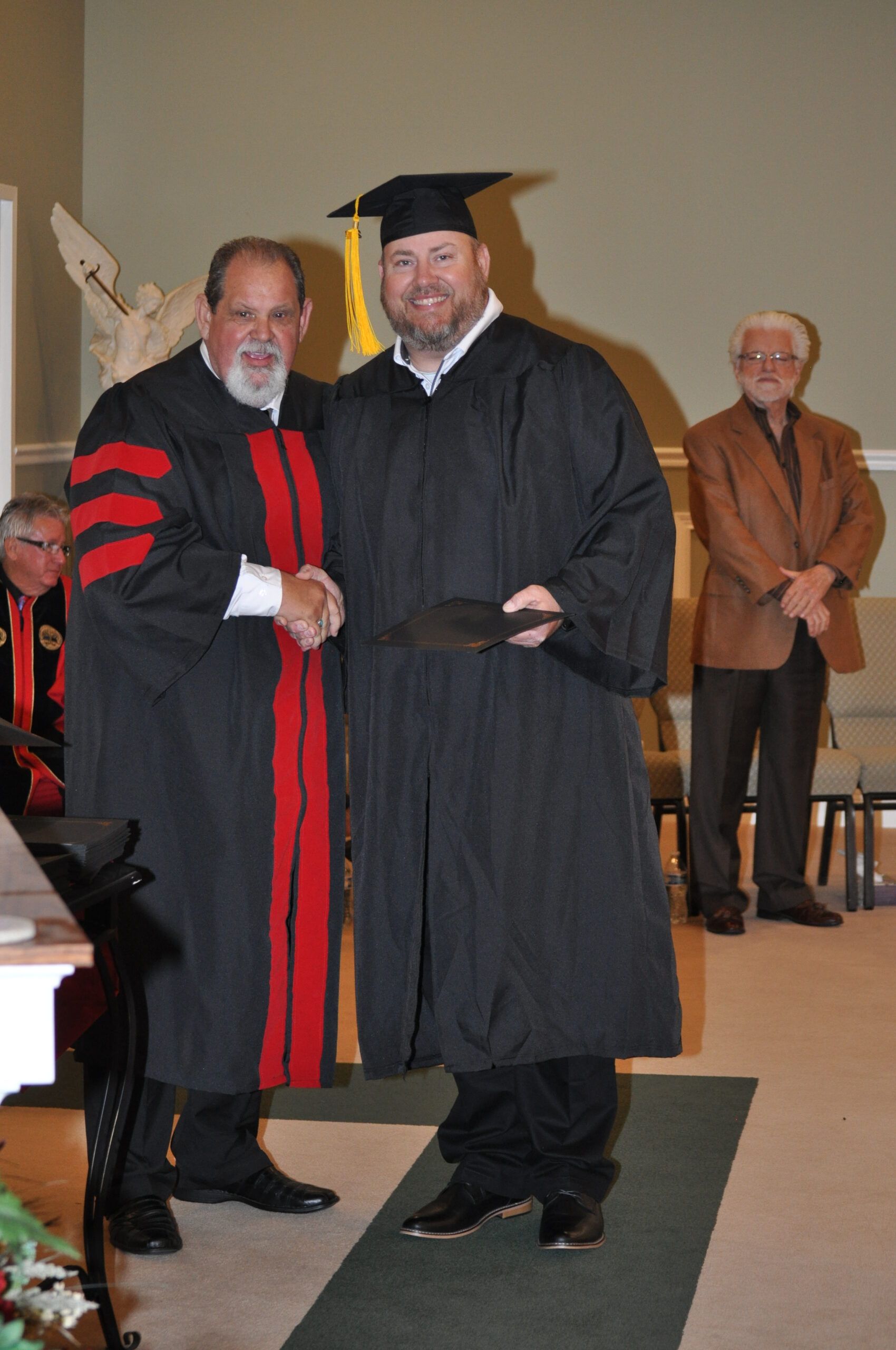 Man in graduation cap and gown receiving diploma, shaking hands with another person in academic attire.