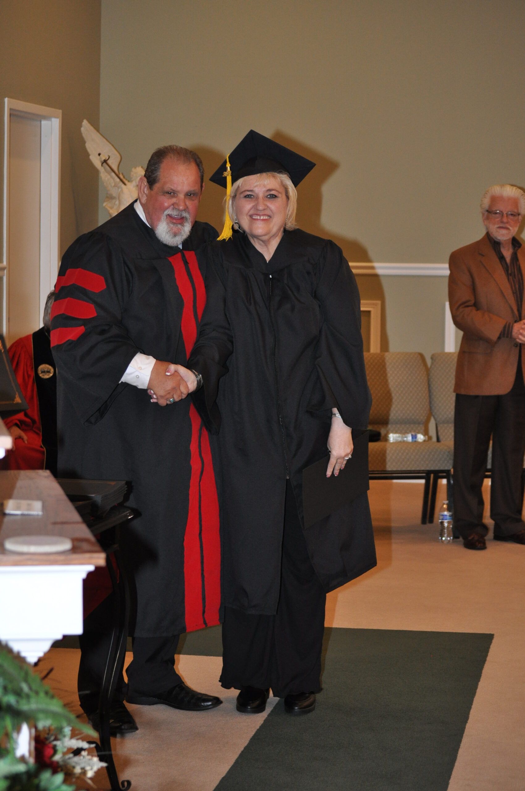 Man in academic regalia shakes hands with a woman in graduation cap and gown; interior setting.