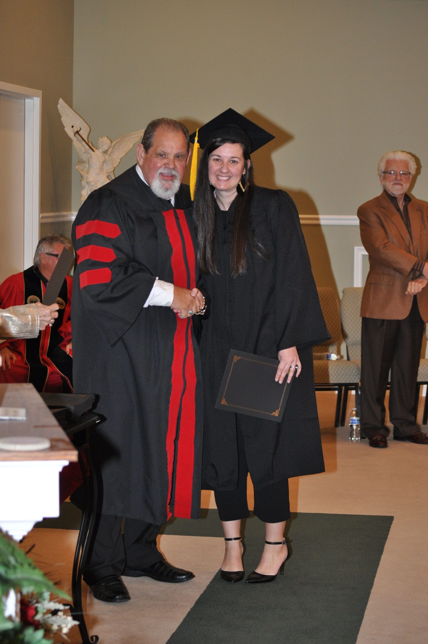 Man in graduation gown shakes hands with a graduate in cap and gown, indoors.