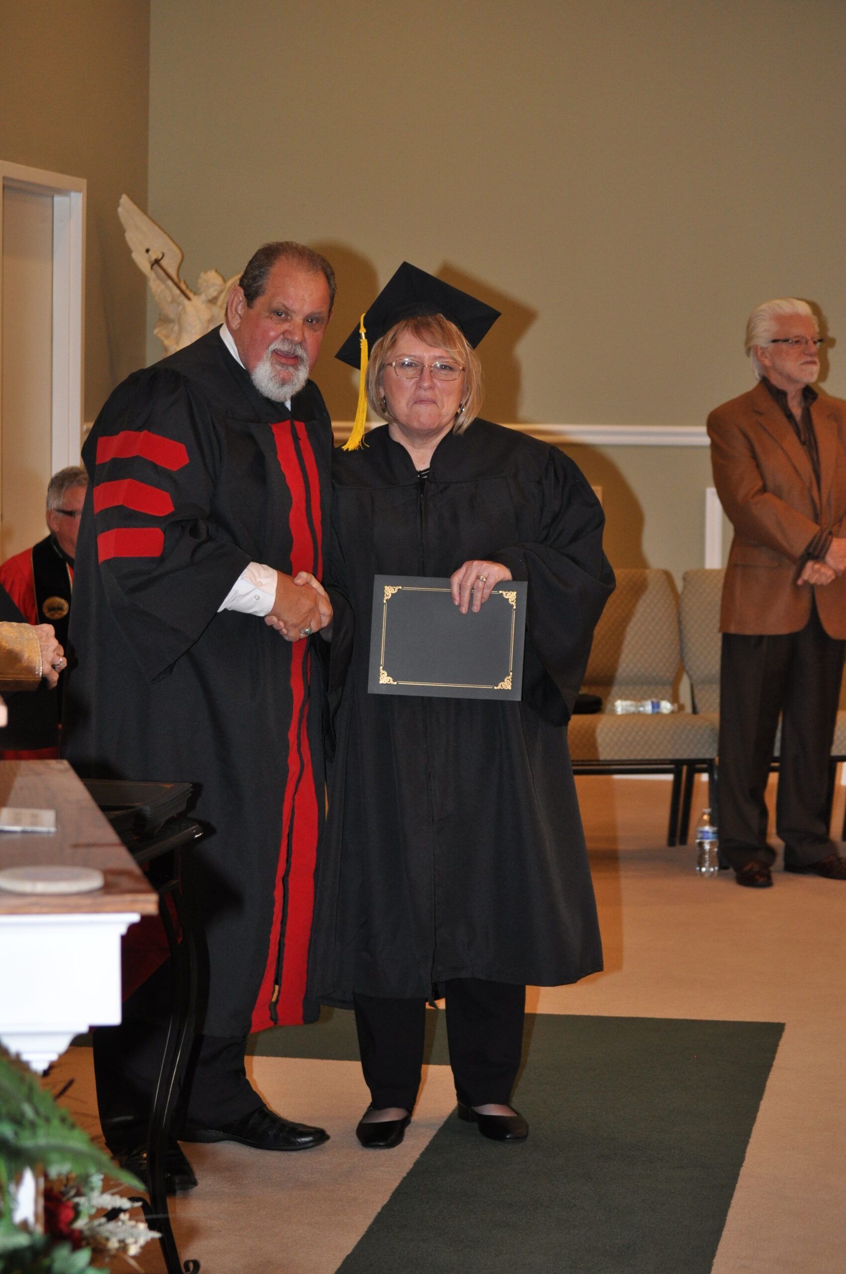 Graduation: Man in academic gown shakes hands with woman in cap and gown, holding a diploma.