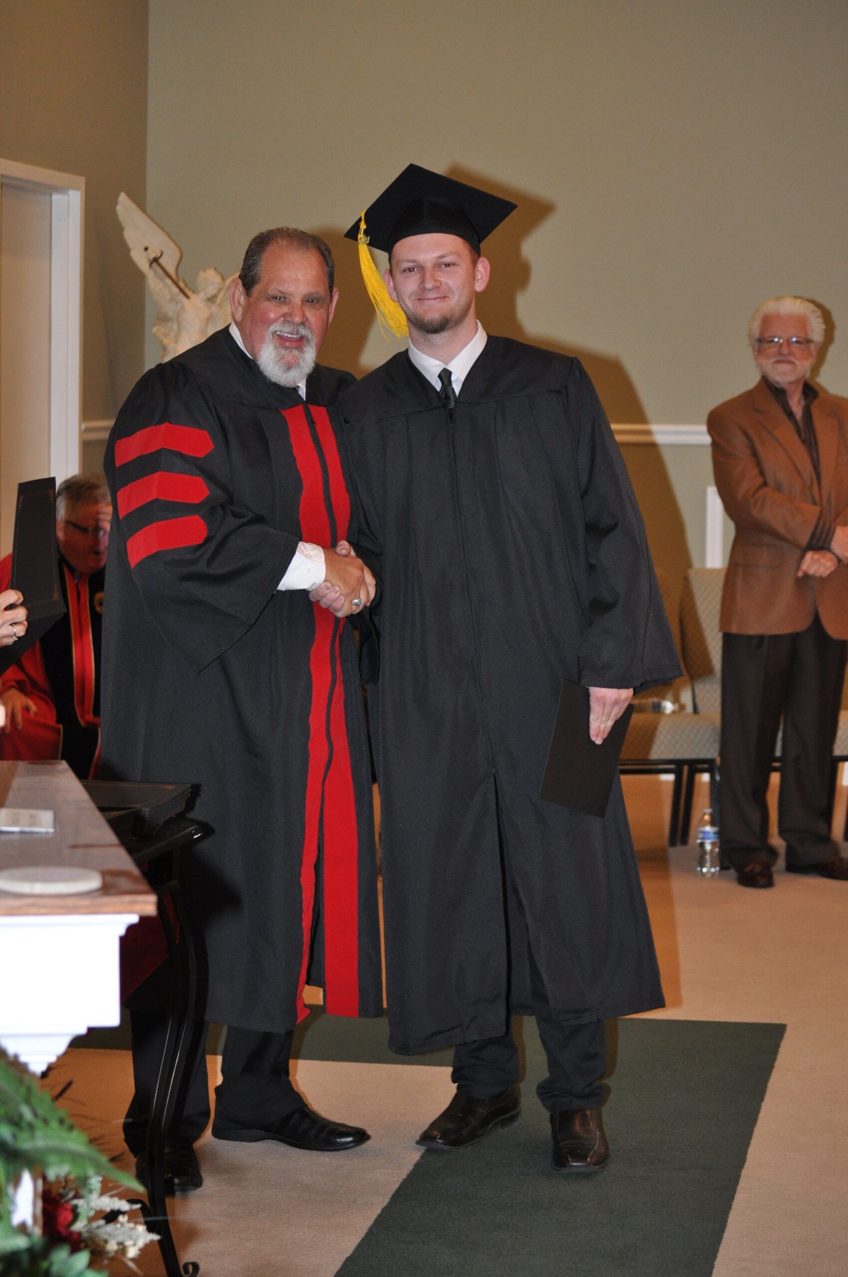 Man in graduation gown shakes hands with robed man, indoors.