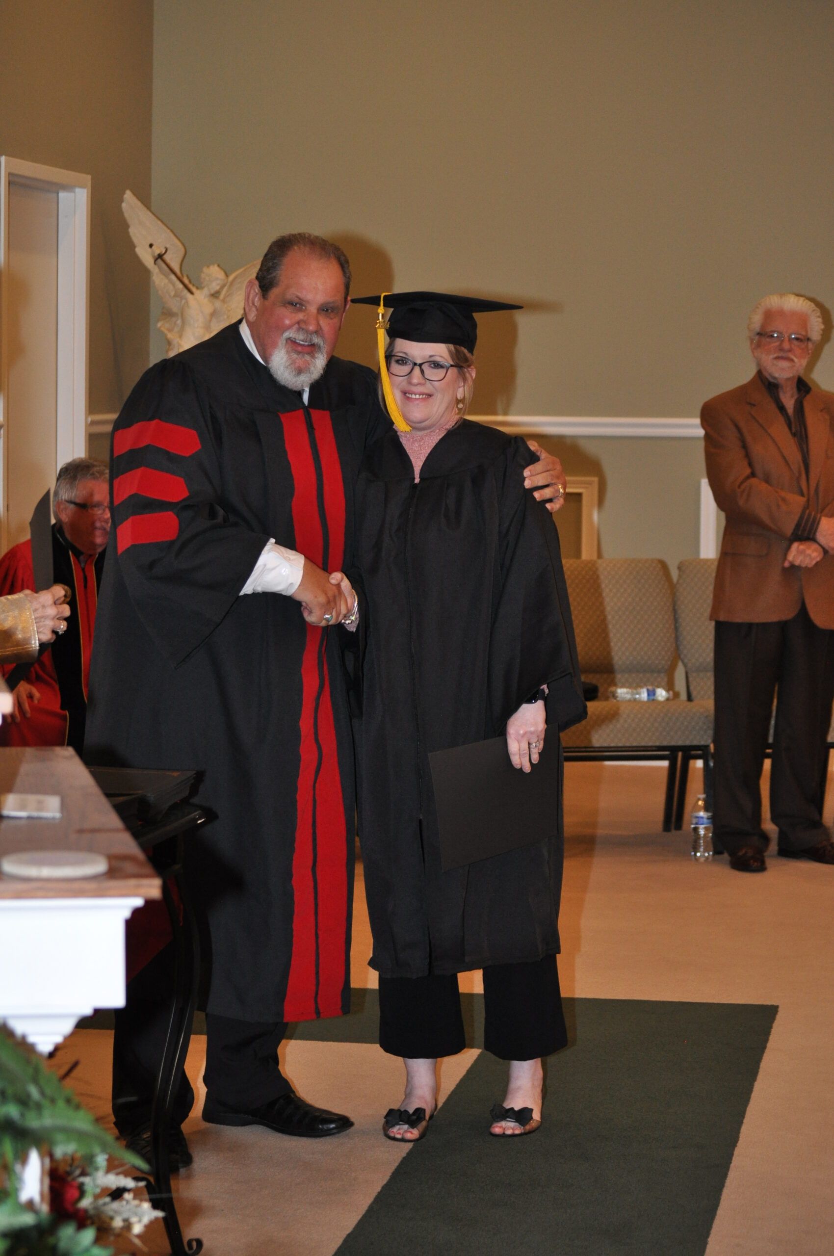 Man in academic robe shakes hands with graduate wearing cap and gown.