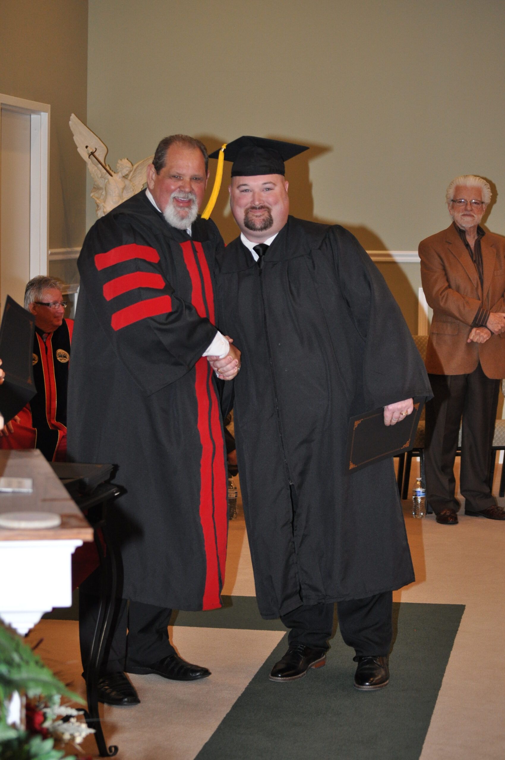 Man in cap and gown shakes hands with a person in academic regalia; graduation ceremony setting.
