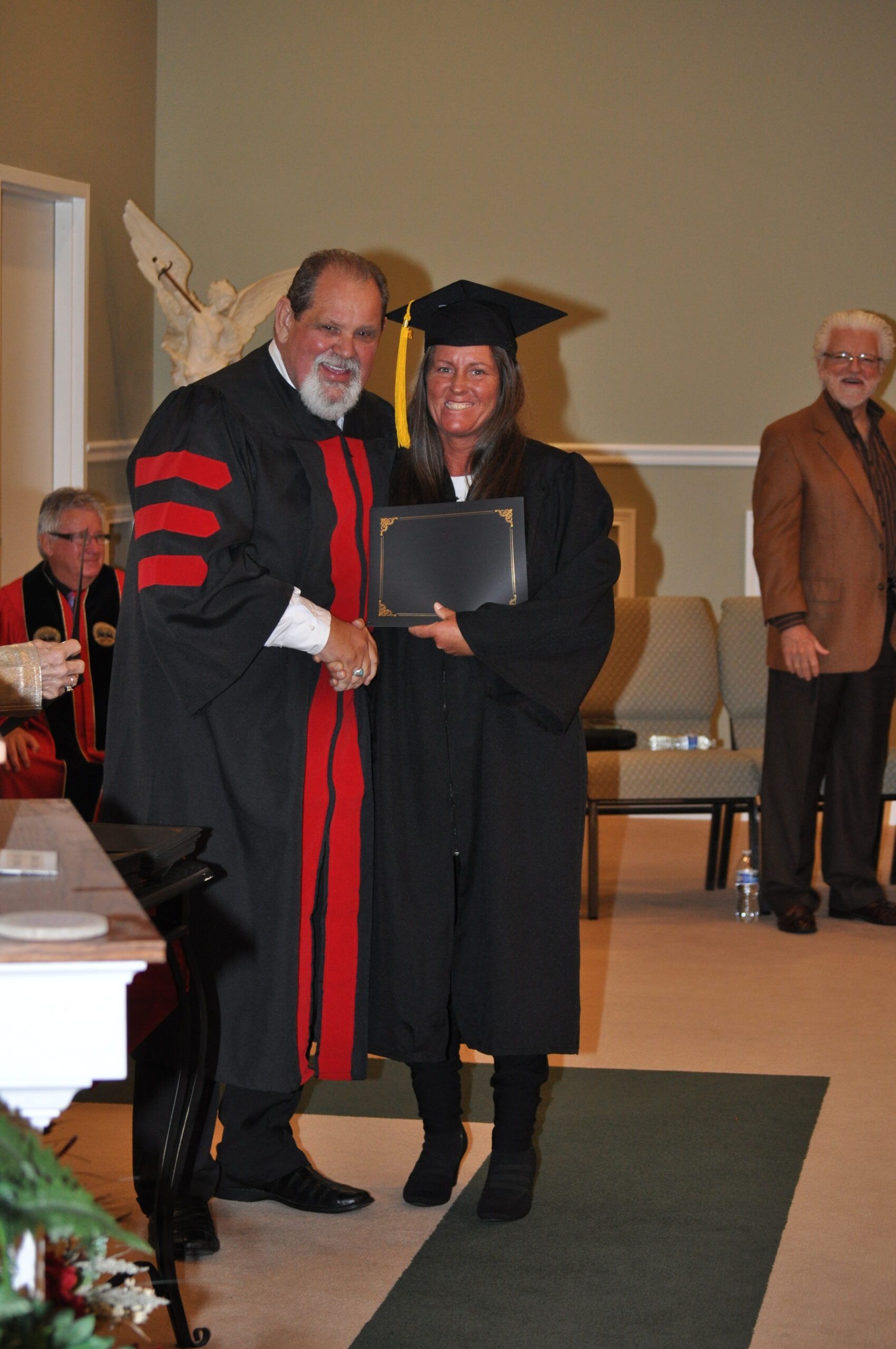 Person in graduation gown receives diploma from robed person, indoors.