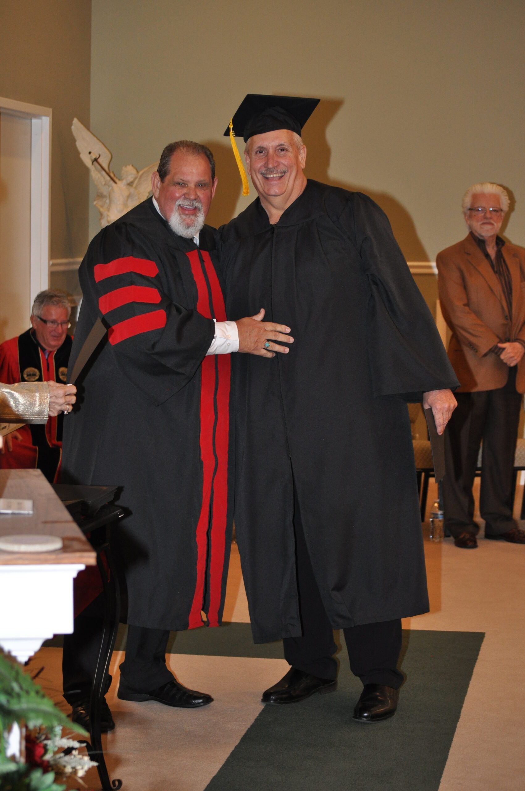 Man in graduation gown receiving an award from another man in robes. Interior setting, smiling.
