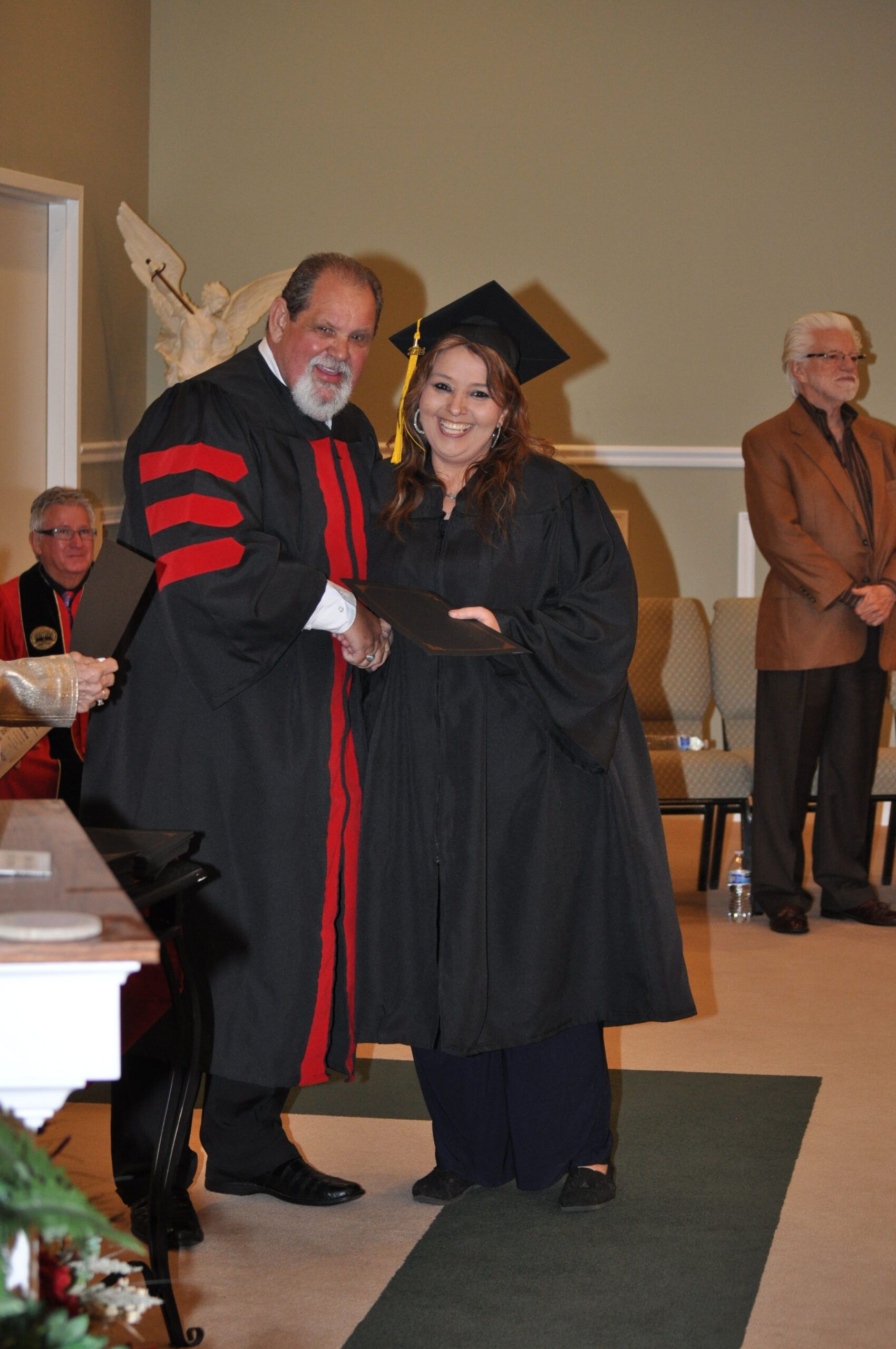 Graduation ceremony: A robed professor shakes hands with a graduate in cap and gown, indoors, other people watching.