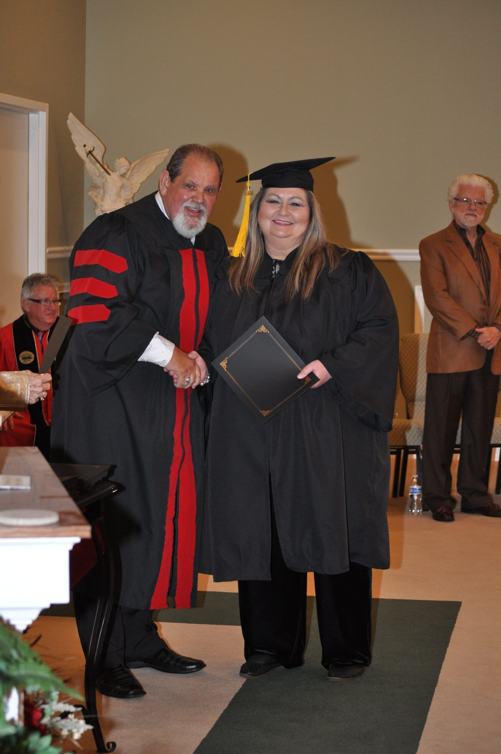 Person in graduation gown shakes hands with person in academic regalia, receiving a diploma.