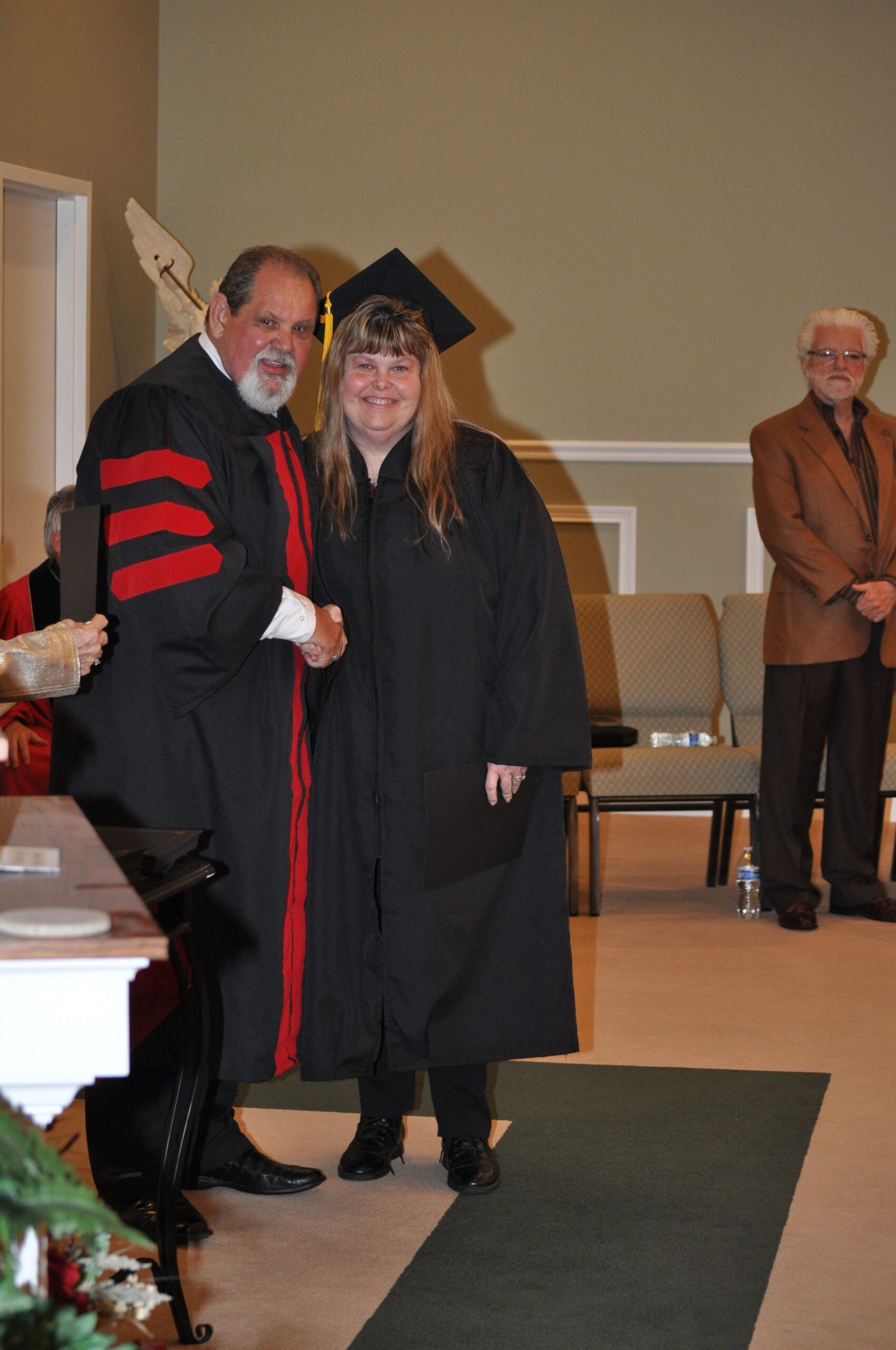 A graduate in a cap and gown shakes hands with a person in academic regalia, inside a building.