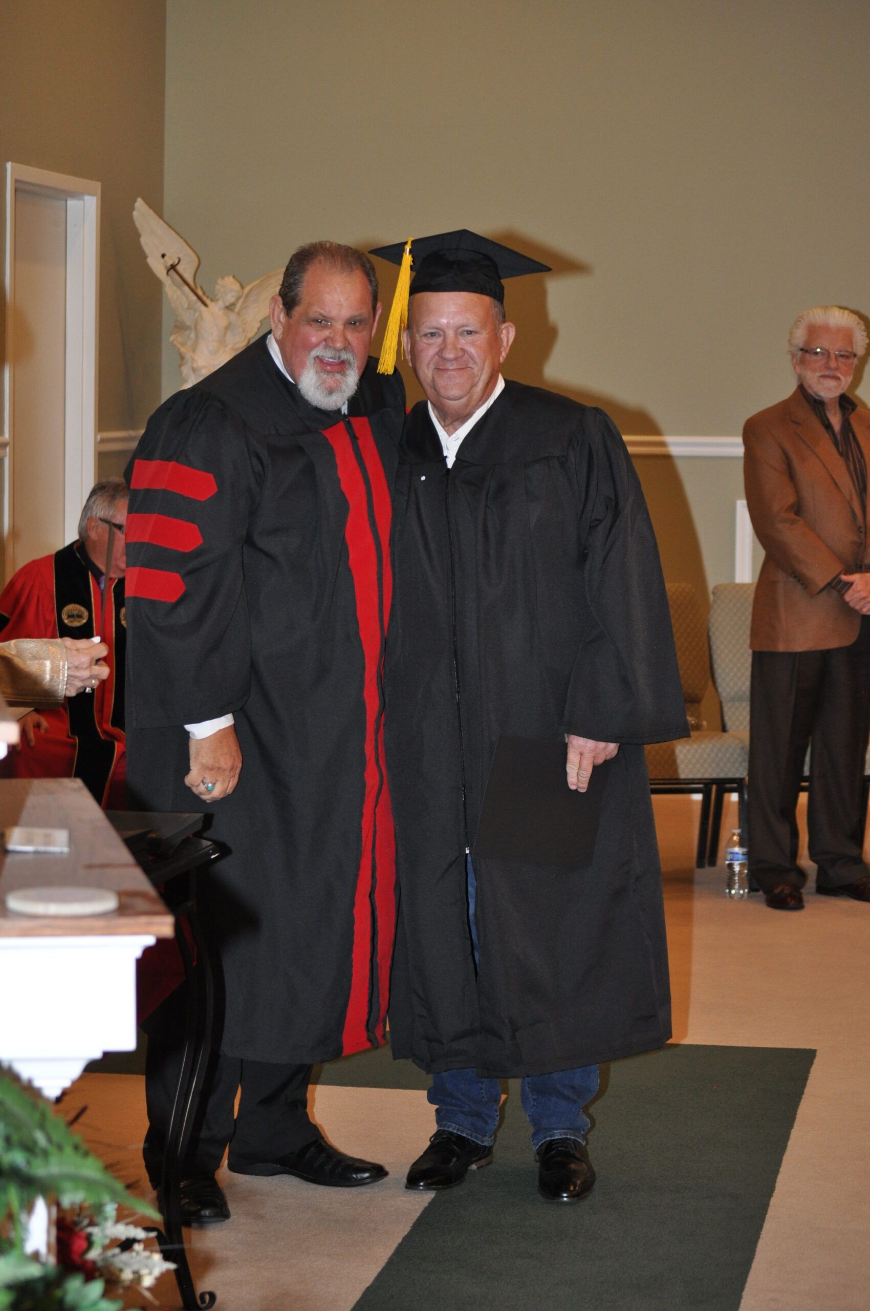 Two men in graduation gowns, one with a mortarboard, pose indoors, smiling.
