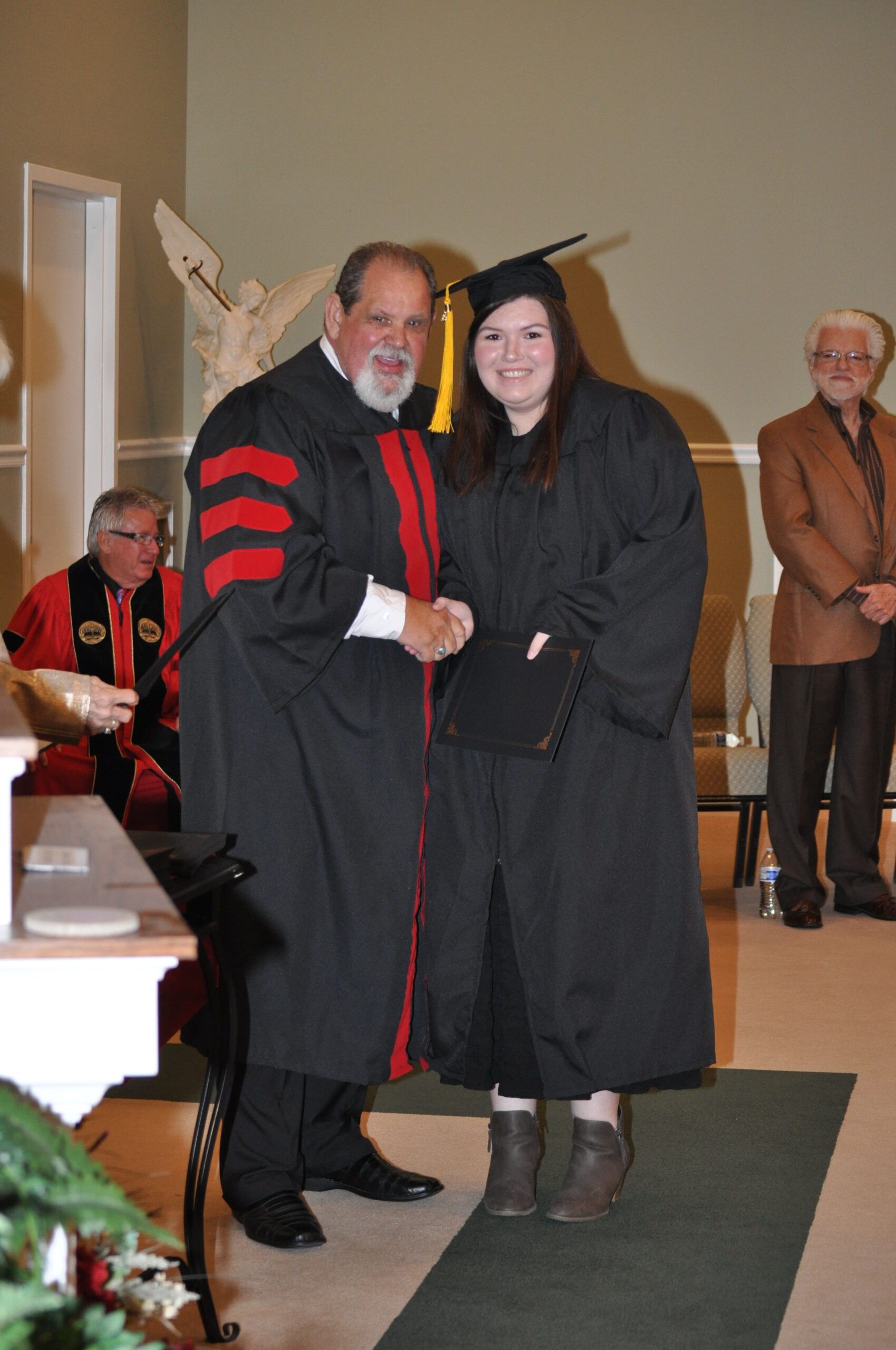 Person in cap and gown shakes hands with a person in academic regalia; graduation ceremony.