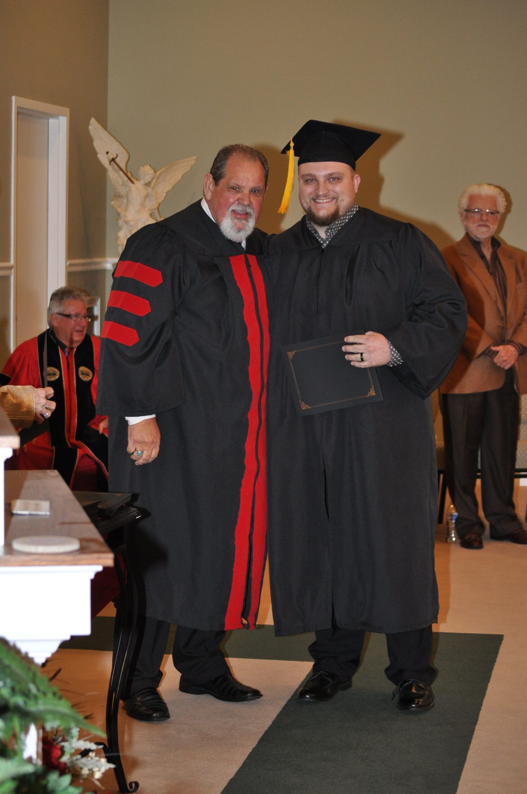 Two men in graduation gowns, one with a mortarboard, pose for a photo in a church.