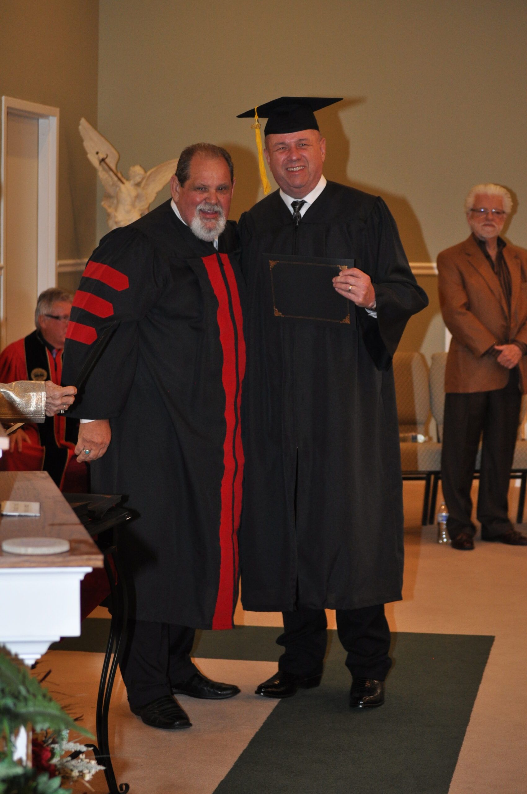 Man in cap and gown receives diploma from a robed figure, indoors on a red carpet.