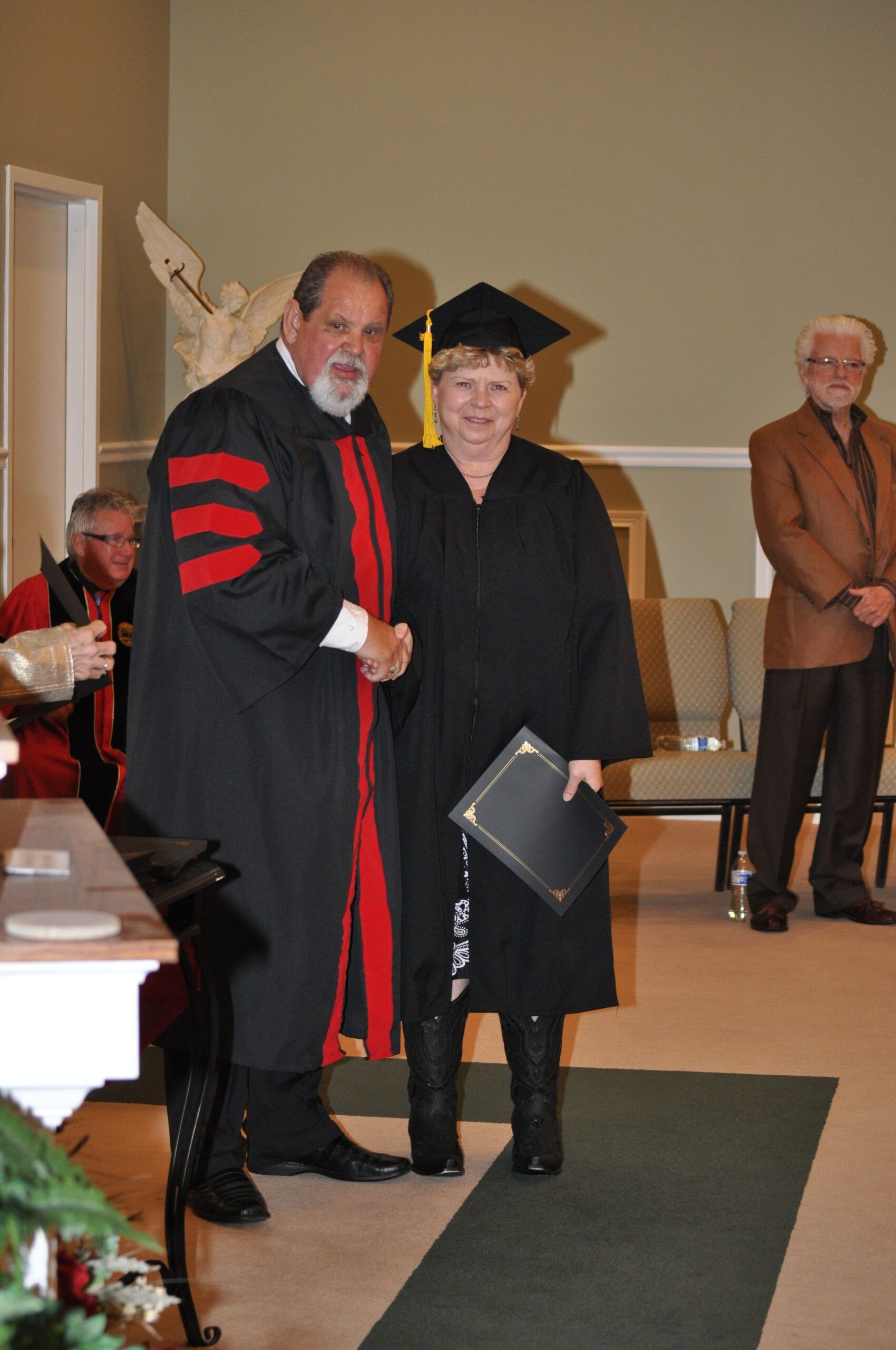 Man in academic robe shakes hands with graduate wearing cap and gown.