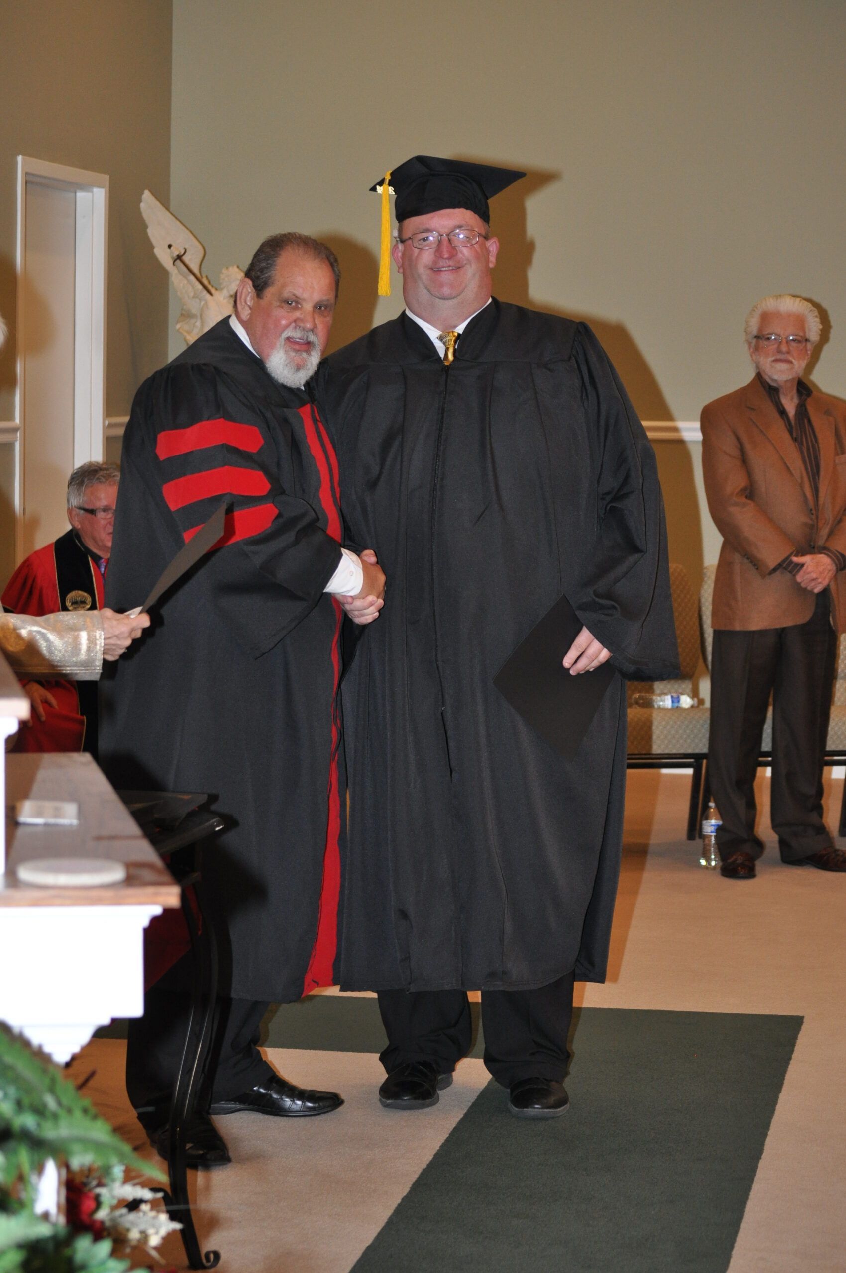 Man in graduation cap and gown shakes hands with a man in academic regalia; indoors.