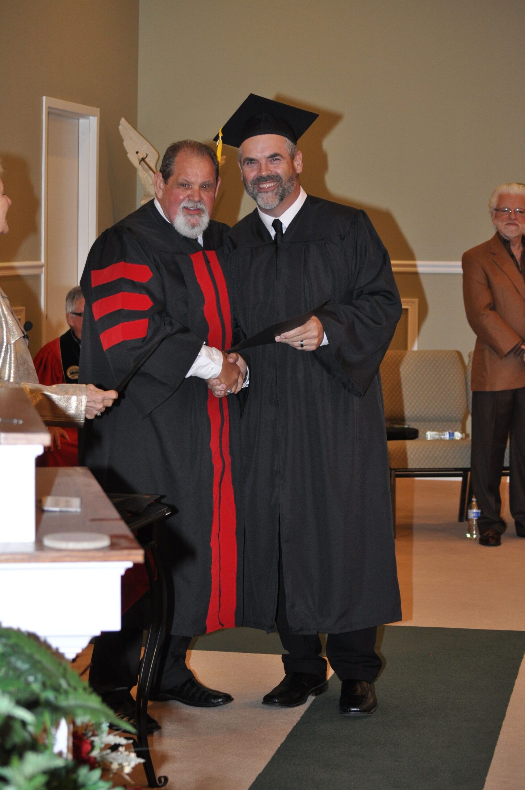 Man in graduation gown receives diploma, shaking hands with another in academic regalia. Indoor ceremony.