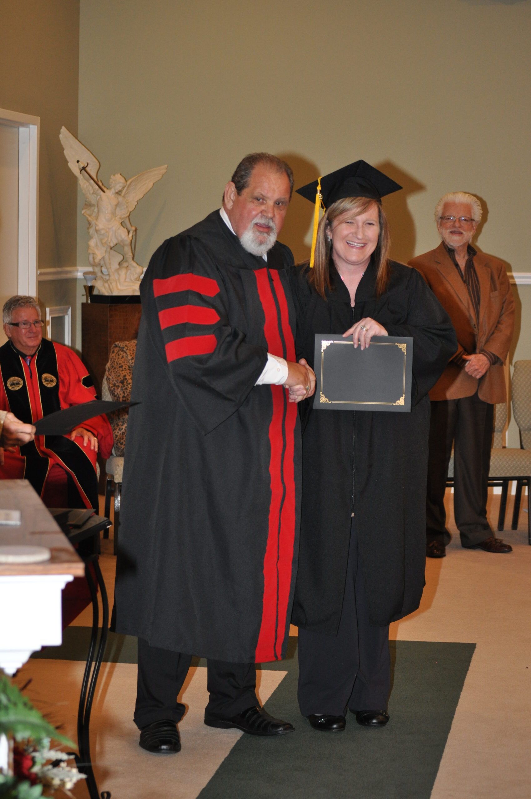 A graduate in black cap and gown shakes hands with a person in a red and black academic robe.