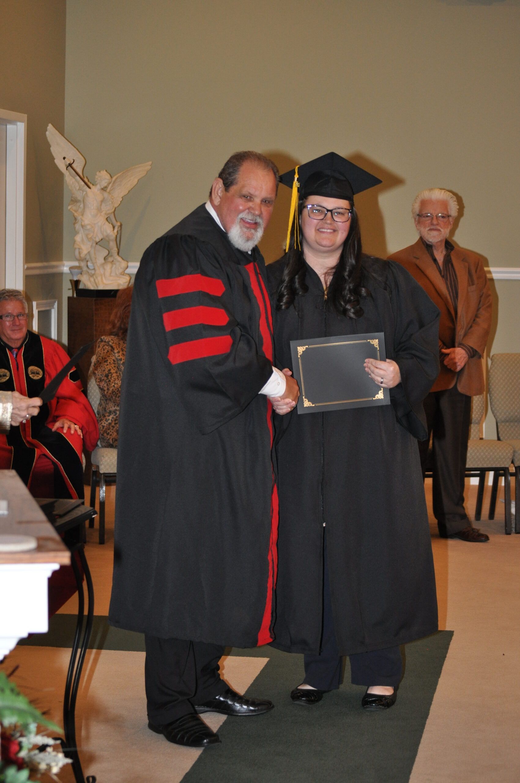 Woman in cap and gown receiving diploma, shaking hands with a man in academic robes at a graduation ceremony.