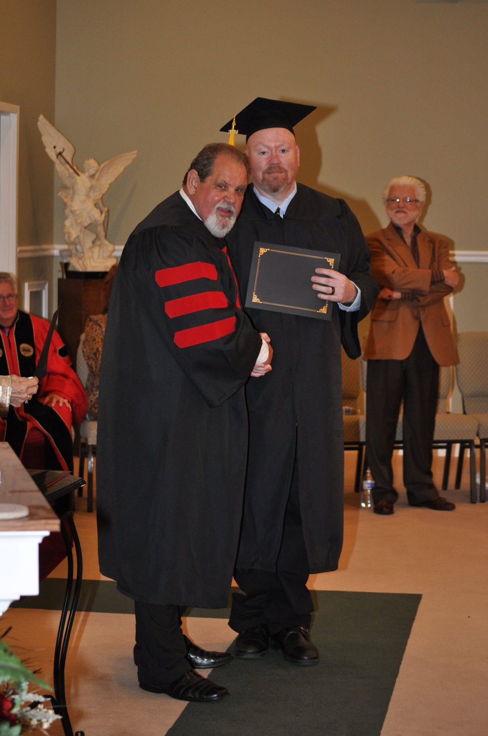Man in graduation gown receiving diploma from another, with onlookers.