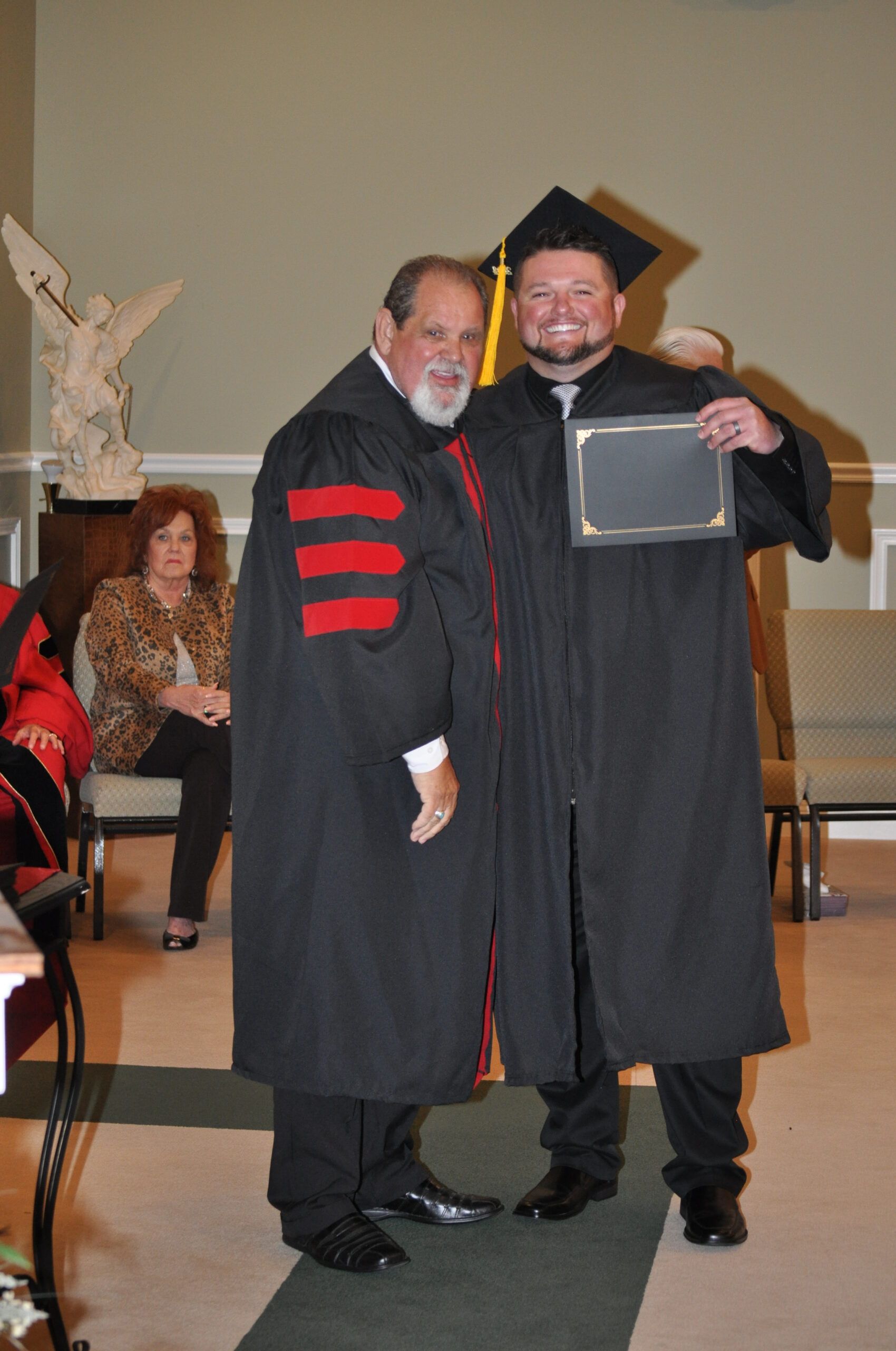 Man in graduation gown receiving a diploma from another man, inside a building.